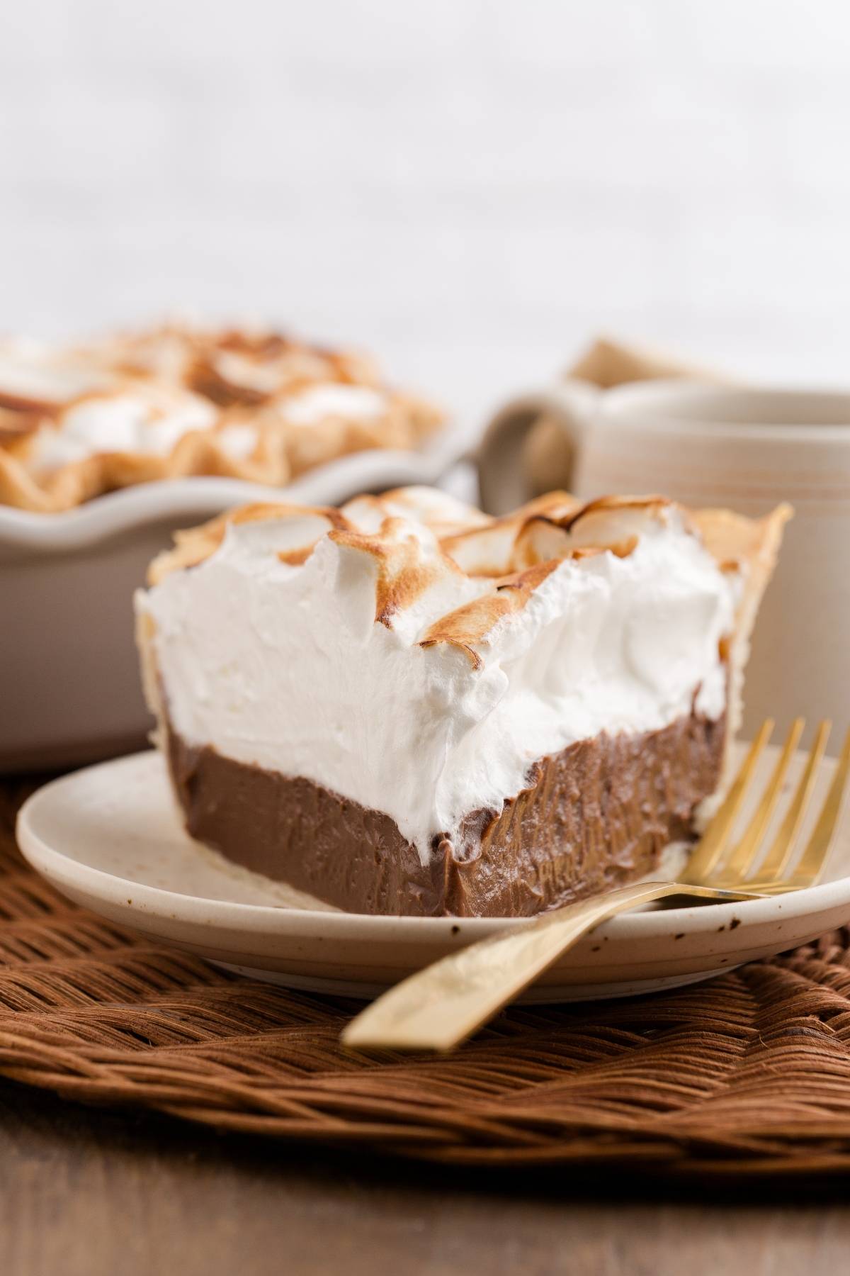A slice of chocolate pie with whipped topping on a plate with a fork, pie dish in the background.