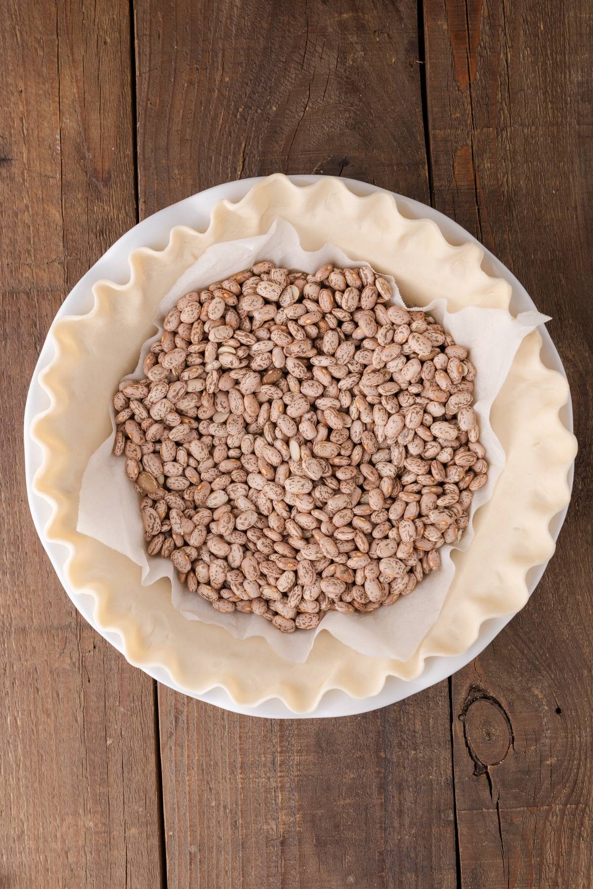 Unbaked pie crust in a white dish, lined with parchment paper and filled with dried beans as pie weights.
