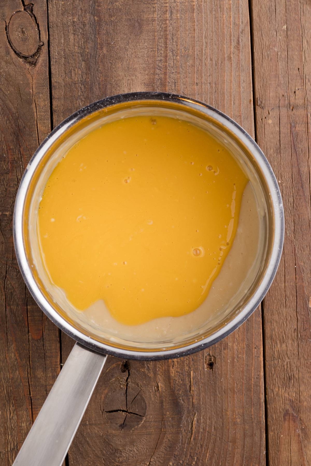 A saucepan with yellow sauce or custard sits on a rustic wooden surface.