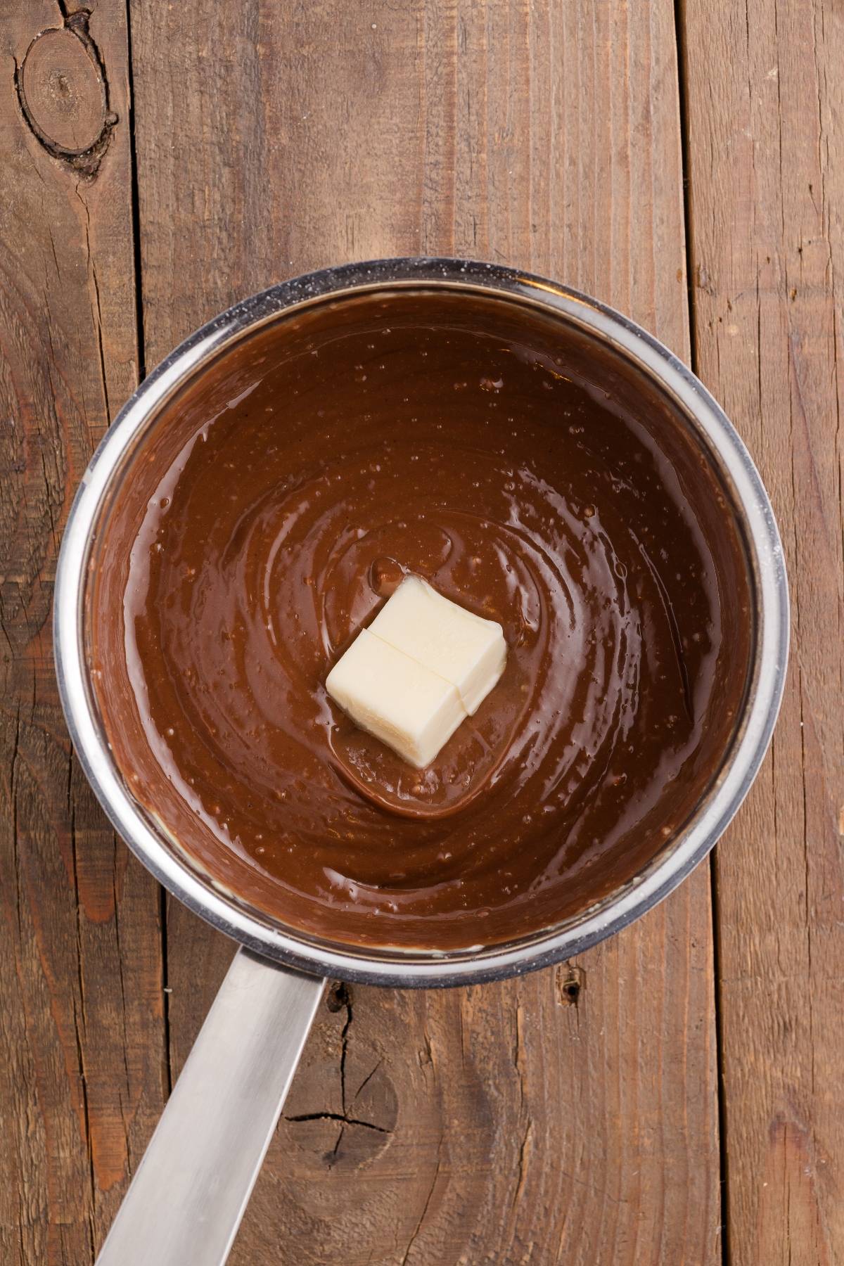 A saucepan of melted chocolate with a pat of butter on top, set on a rustic wooden surface.
