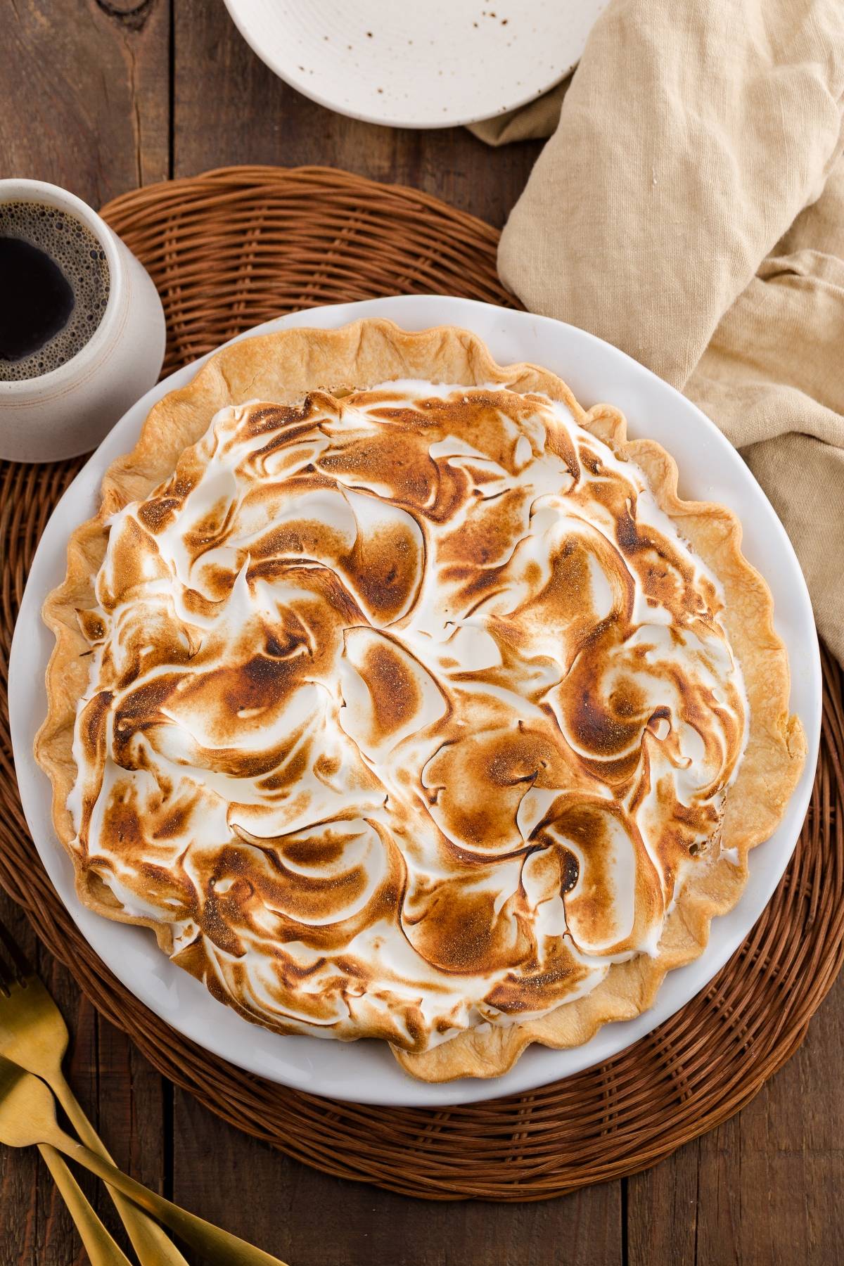 A pie with toasted meringue topping on a white plate, next to a cup of coffee and gold utensils.