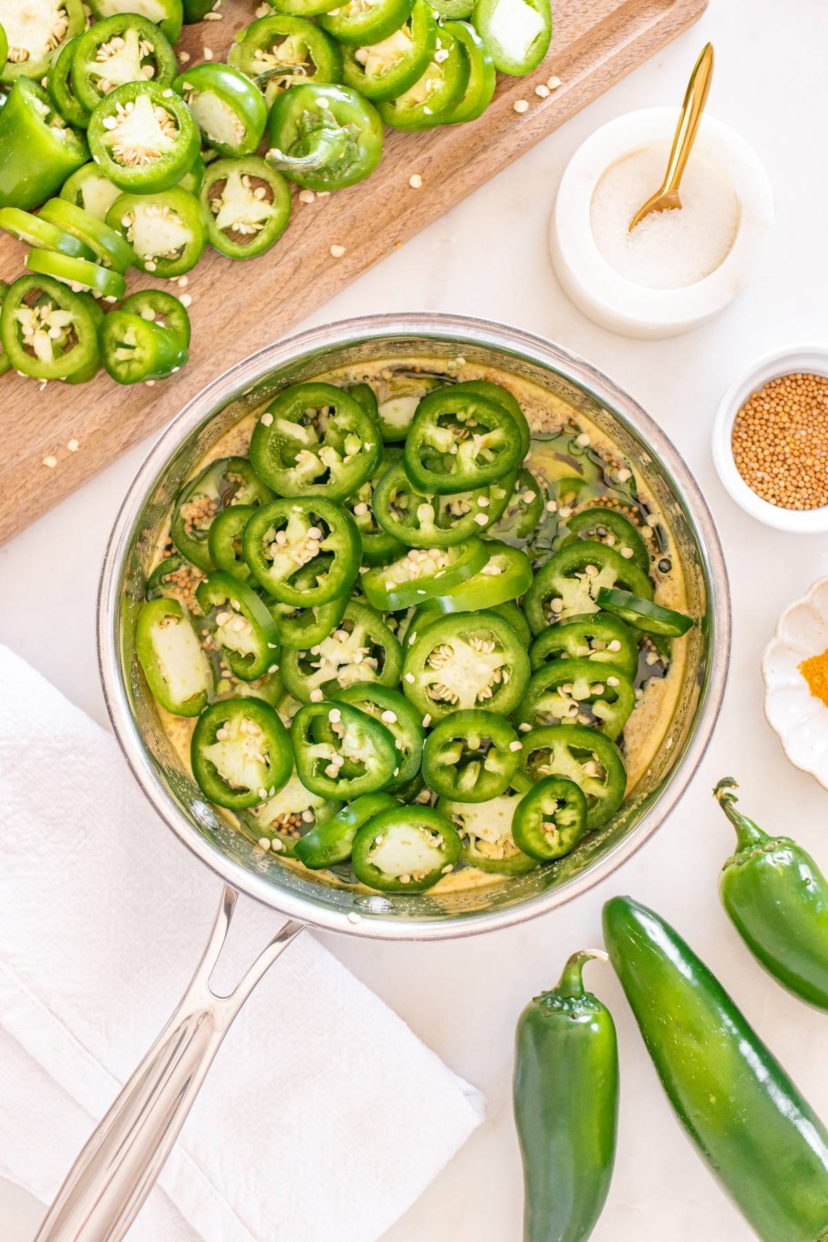 Sliced jalapeños in a saucepan with pickling spices, surrounded by whole jalapeños and seasoning bowls.