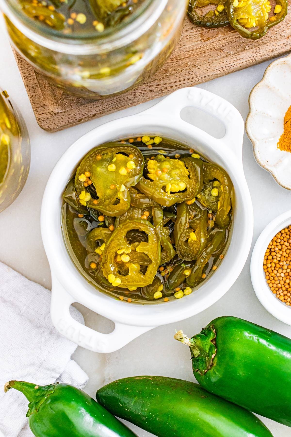 A white bowl filled with pickled jalapeño slices, surrounded by fresh jalapeños and spices.