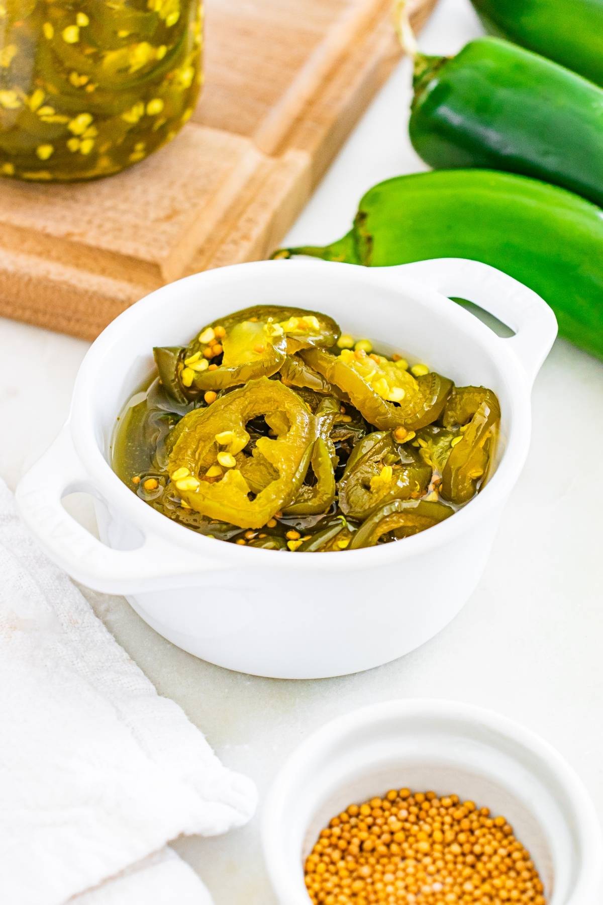 Sliced pickled jalapeños in a white bowl, with fresh jalapeños and mustard seeds nearby.