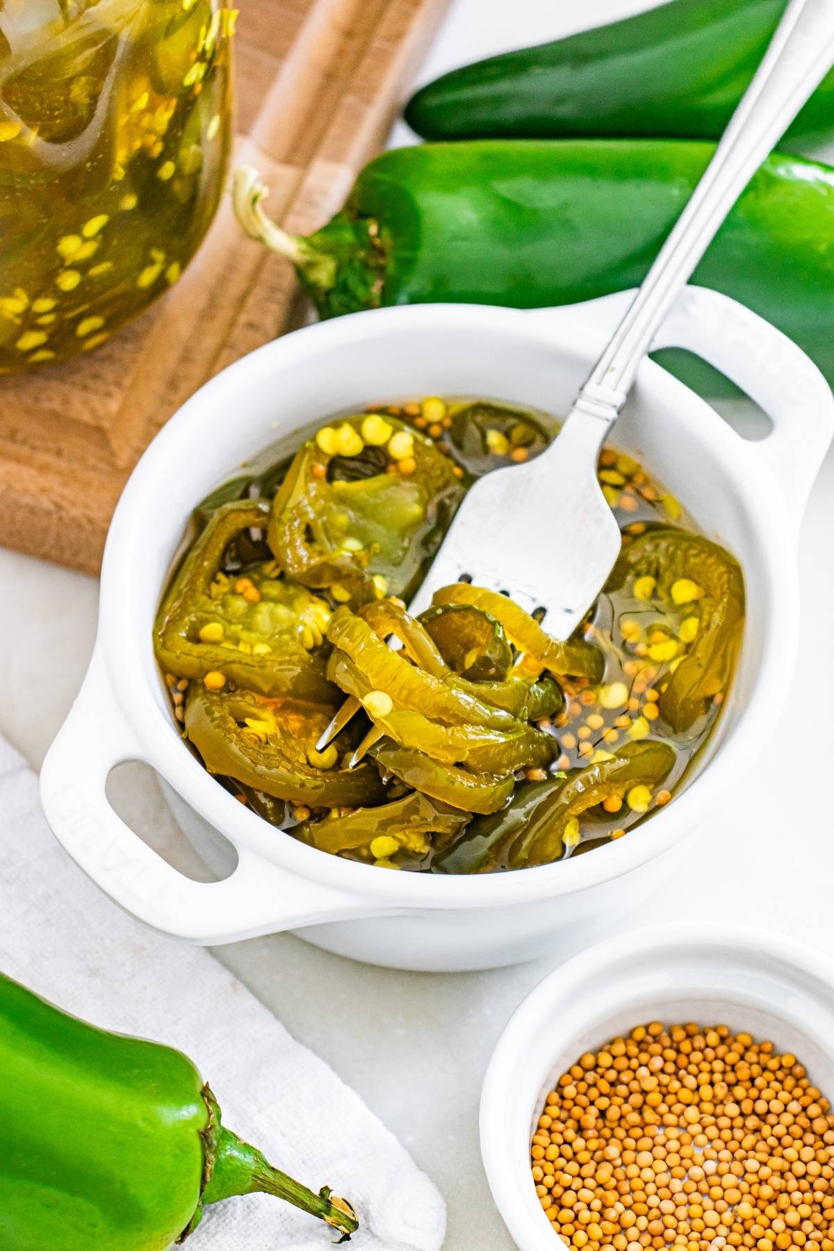 A white bowl with pickled jalapeño slices, a fork, fresh jalapeños, and mustard seeds nearby.
