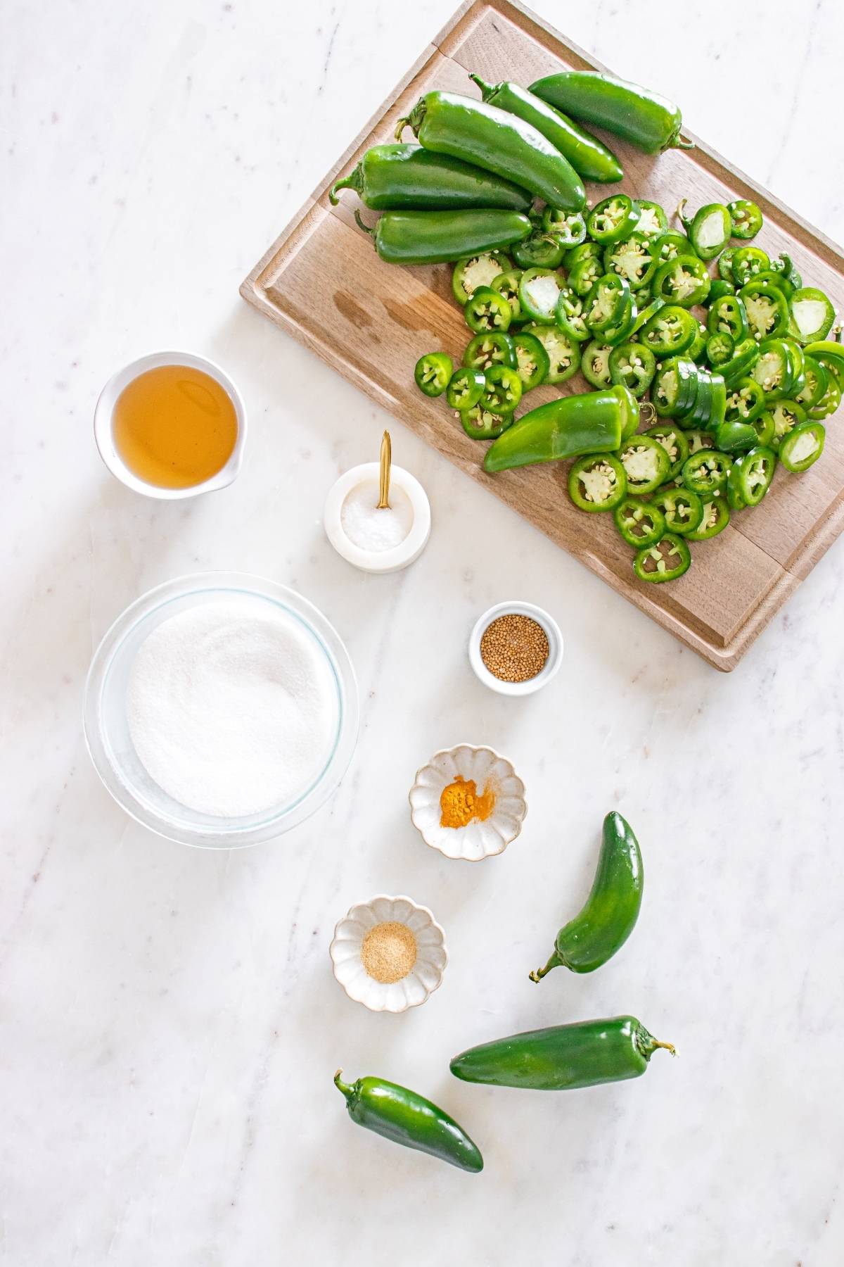 Sliced and whole jalapeños, sugar, honey, spices, and salt arranged on a white surface with a cutting board.