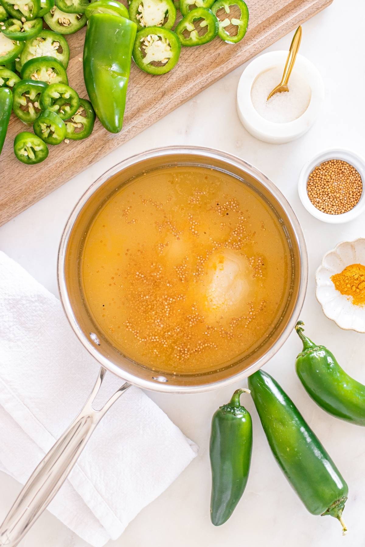 Pot with yellow liquid and mustard seeds, surrounded by jalapeños, salt, turmeric, and a white towel.