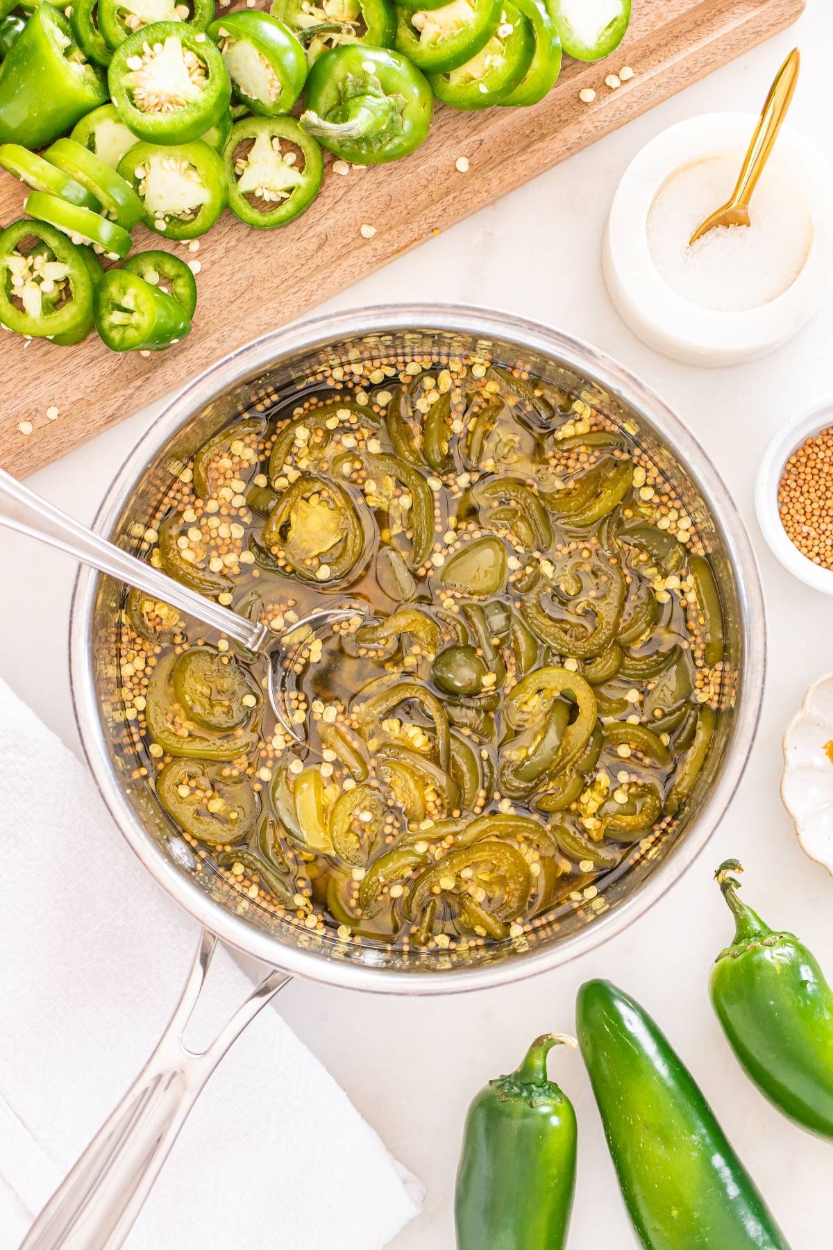 Sliced jalapeños in a saucepan with pickling spices, next to fresh jalapeños and a bowl of salt.