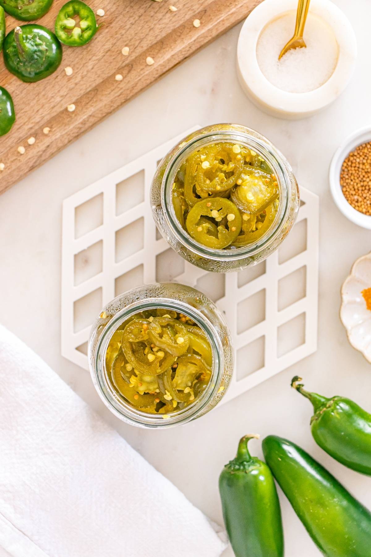 Two jars filled with pickled jalapeño slices on a counter, surrounded by fresh jalapeños and spices.