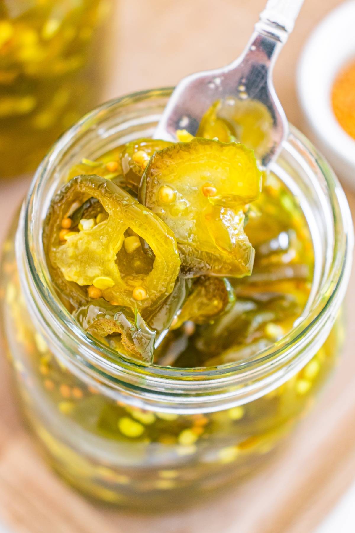 A fork lifts candied jalapeño slices from a glass jar filled with pickled jalapeños and seeds.