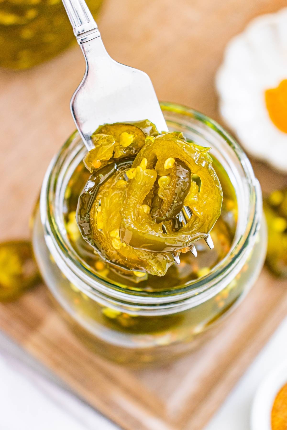 A fork lifts sliced pickled jalapeños from a glass jar on a wooden surface.