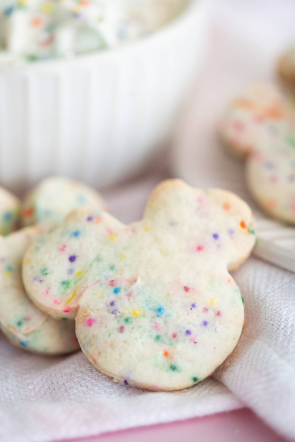 Funfetti bunny-shaped cookies on a white cloth, with colorful sprinkles and a bowl of cookies and cream cheese dip in the background.