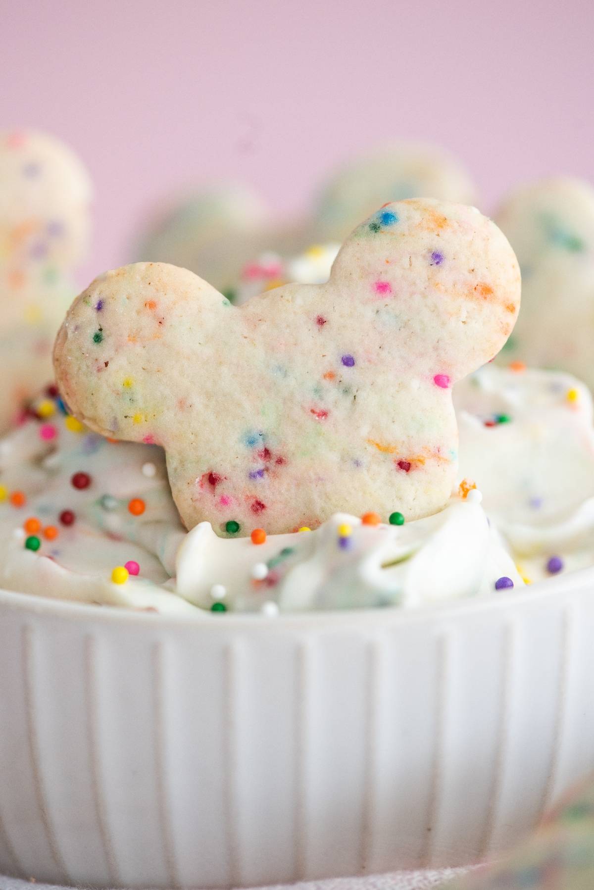 A Mickey Mouse-shaped funfetti cookie dipped in creamy Funfetti Dunkaroo dip with colorful sprinkles, served in a white bowl.