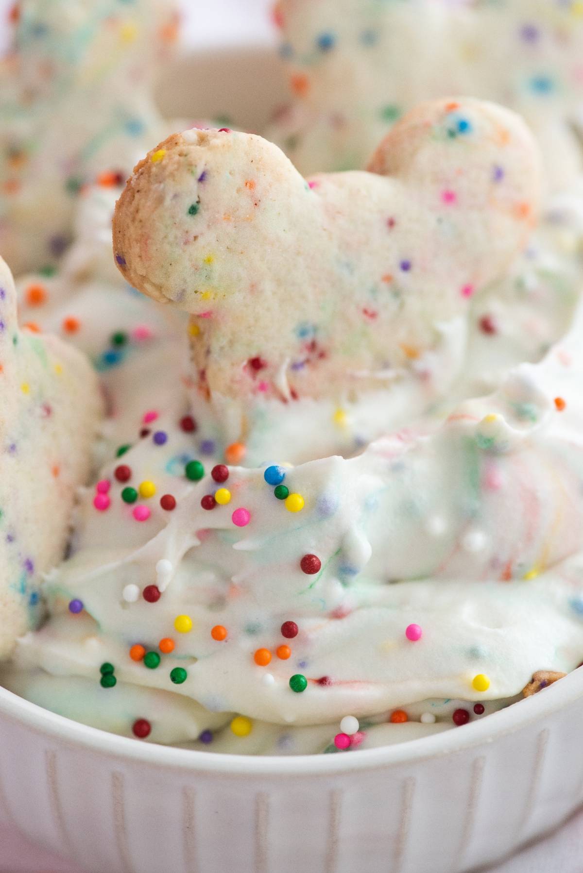 A Mickey Mouse sugar cookie with rainbow sprinkles sits on white frosting, perfect for dipping into Funfetti Dunkaroo dip topped with colorful sprinkles.