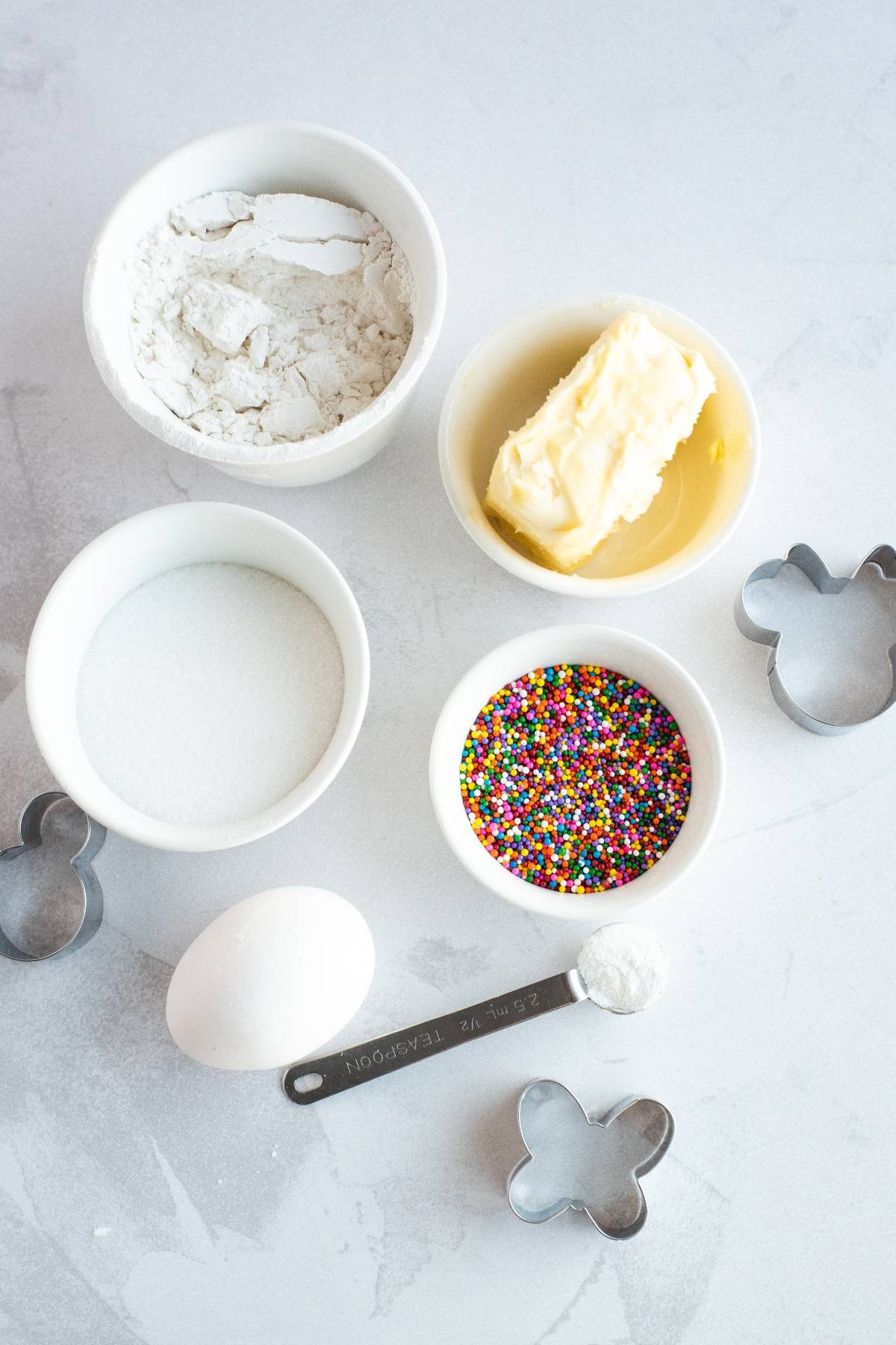 Baking ingredients and cookie cutters on a table, ready to make Mickey Mouse sugar cookies or Disney Sugar Cookies with Dip, featuring flour, sugar, butter, egg, sprinkles, and baking powder.