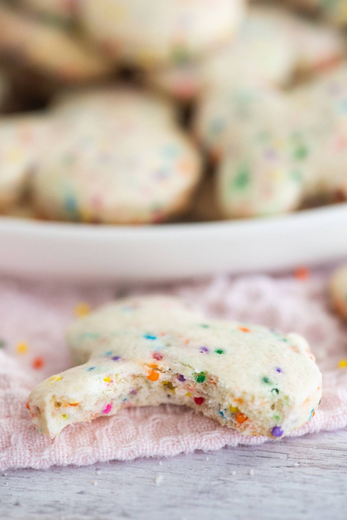 A bitten sprinkle cookie rests on a pink cloth, with more cookies blurred in the background—perfect for pairing with a creamy Funfetti Dunkaroo dip.