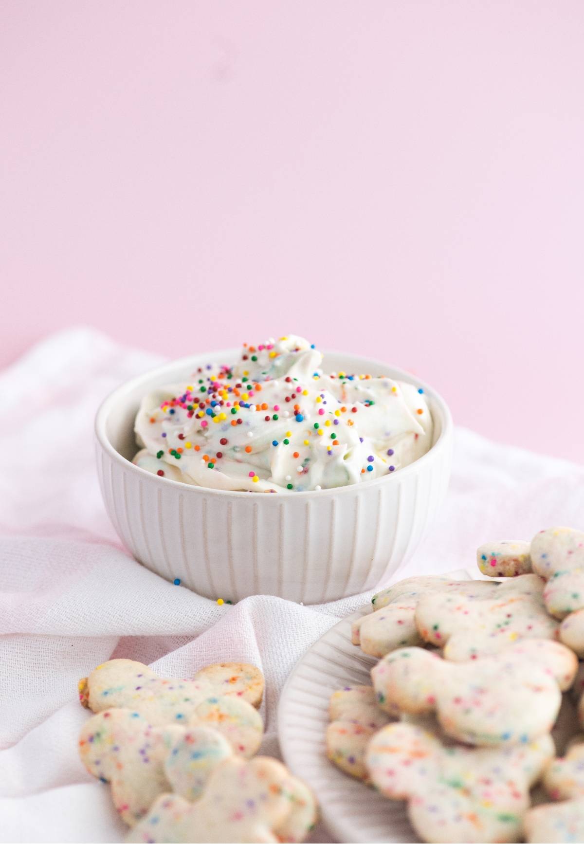 A bowl of Funfetti Dunkaroo dip with rainbow sprinkles sits beside colorful sprinkle cookies, all set on a light pink background.