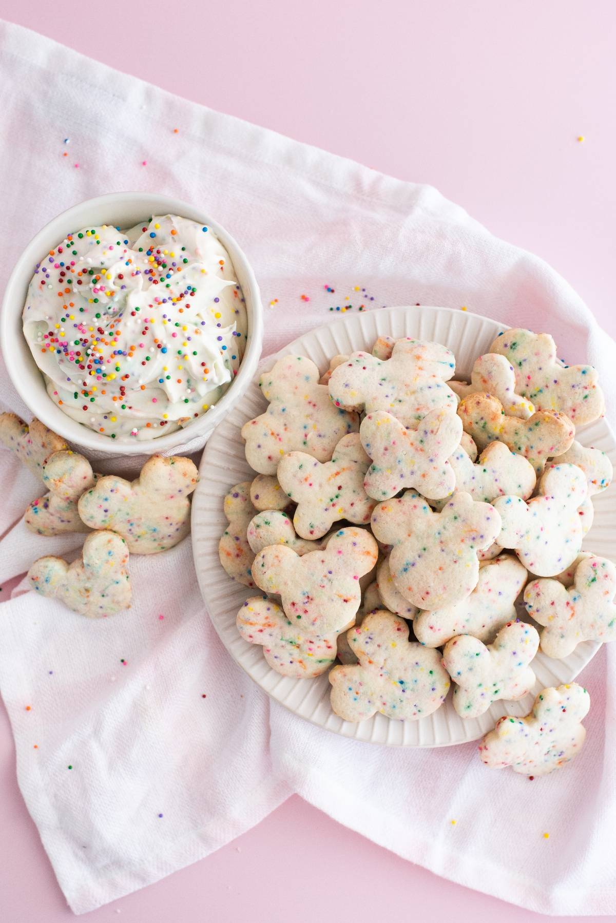 Plate of sprinkle-filled animal-shaped cookies, including Mickey Mouse sugar cookies, with a bowl of cream cheese sprinkle dip topped with rainbow sprinkles, all set on a pink background.
