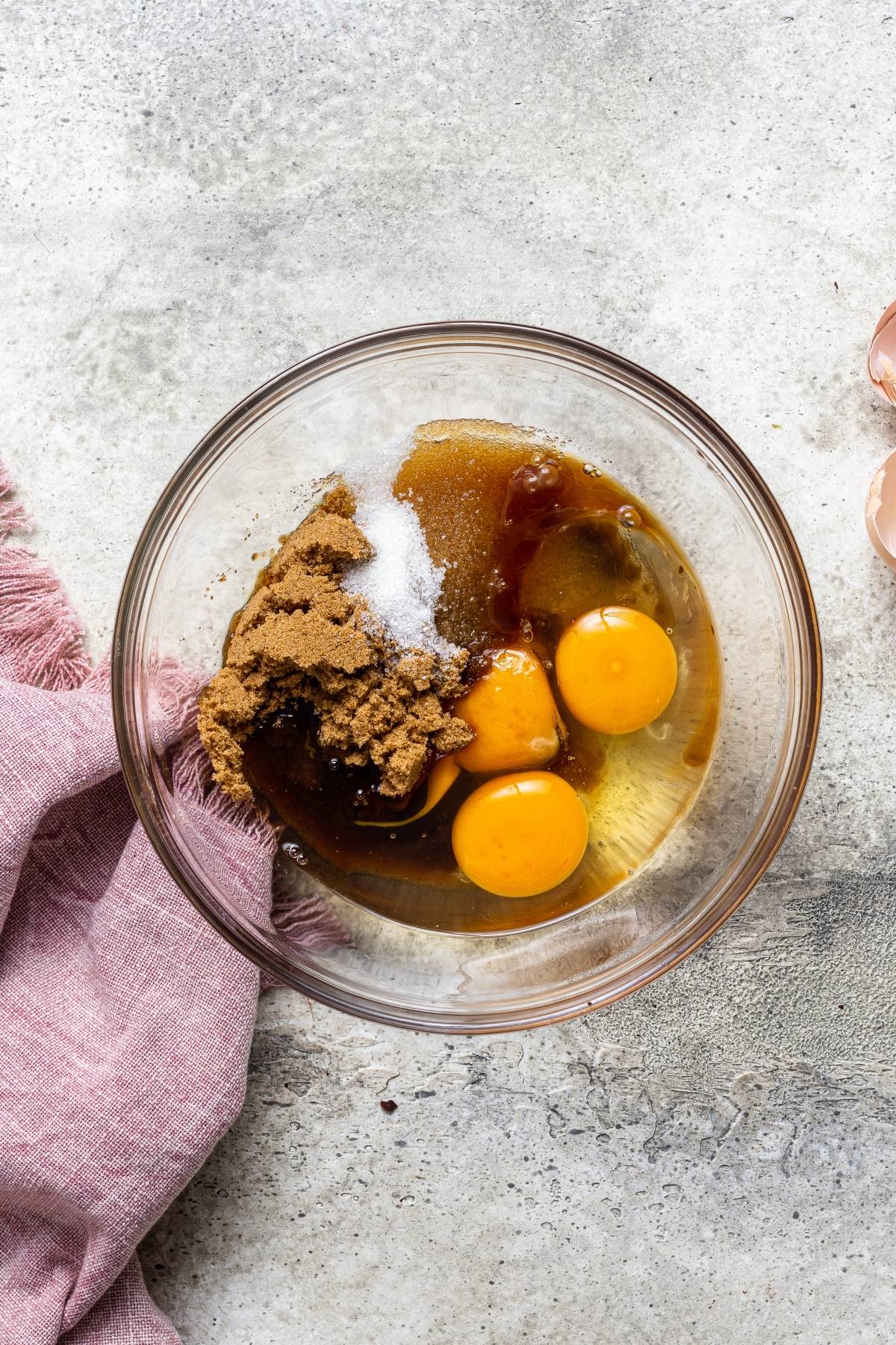 A glass bowl with eggs, brown sugar, vanilla, and salt sits on a gray surface next to a pink cloth—perfect ingredients for easy cherry dessert recipes like Fresh Cherry Brownies.