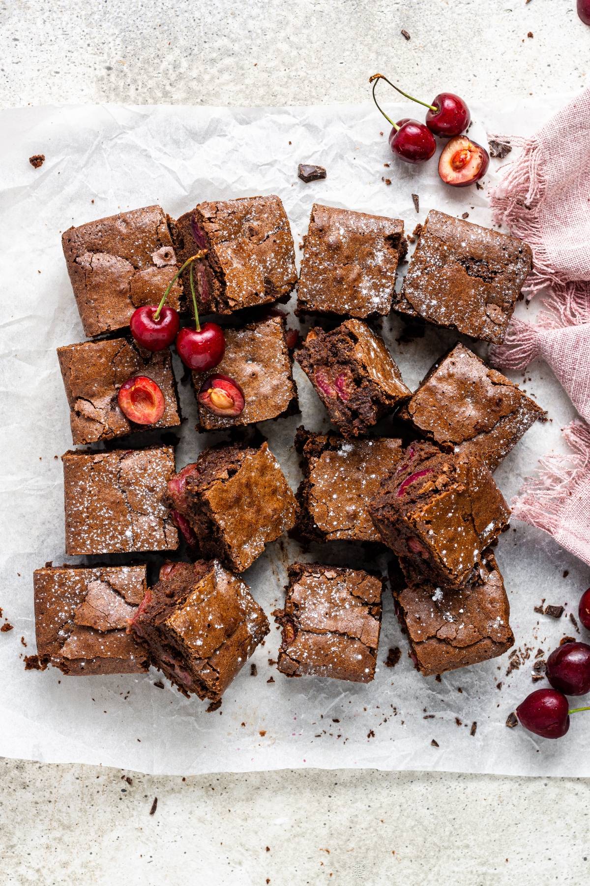 Squares of Double Chocolate Cherry Brownies with cherries on parchment paper, dusted with powdered sugar, and fresh cherries on top—a tempting chocolate and cherry dessert.