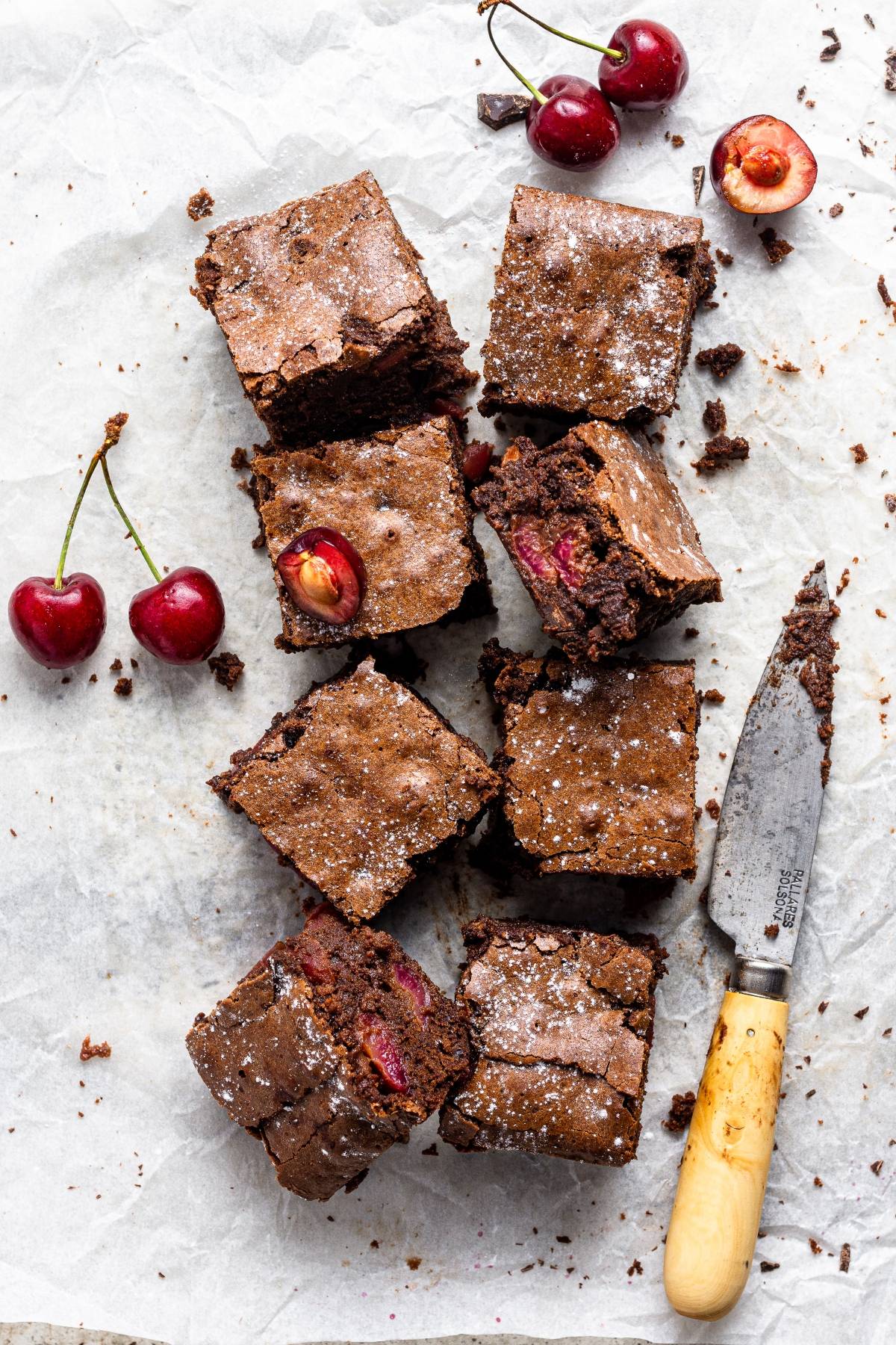 Eight Double Chocolate Cherry Brownies with fresh cherries and a knife rest on parchment paper; some brownies feature cherry pieces inside—an easy cherry dessert perfect for any occasion.