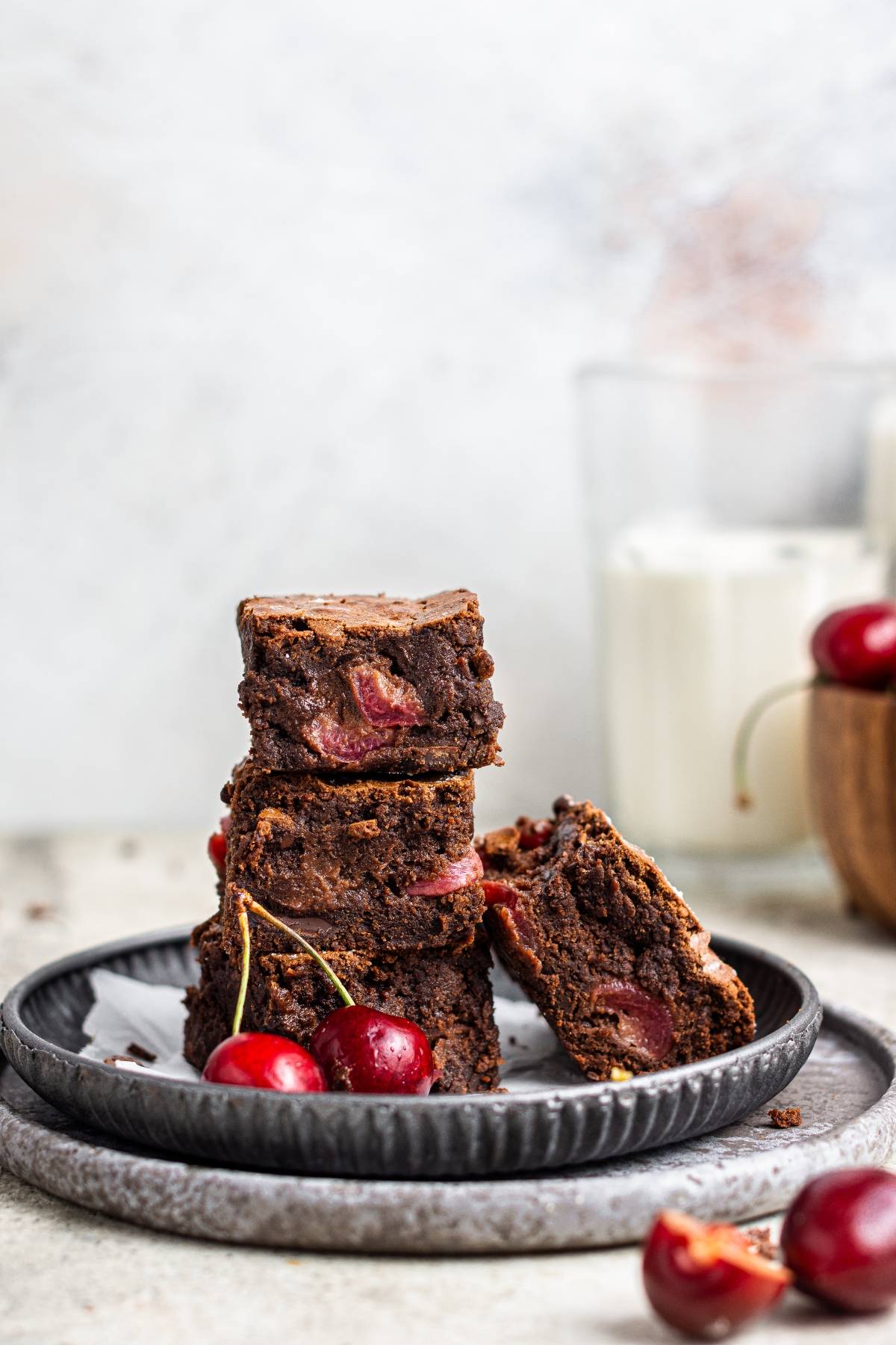 A stack of Fresh cherry brownies with cherries on a black plate, with fresh cherries and a glass of milk in the background—a delicious chocolate and cherry dessert.