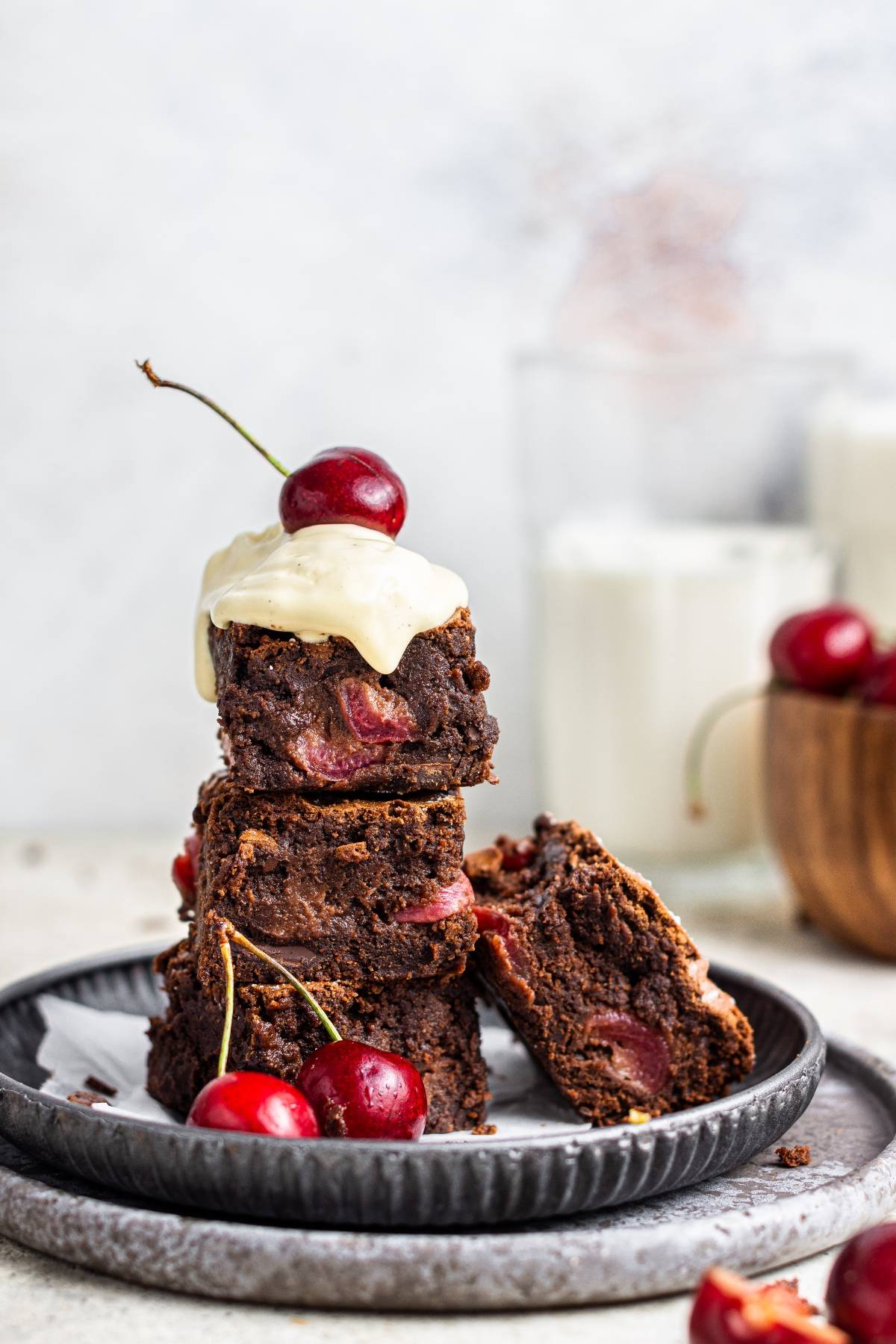 Three fresh cherry brownies stacked, topped with icing and a cherry—this chocolate and cherry dessert is served with extra cherries and milk in the background.