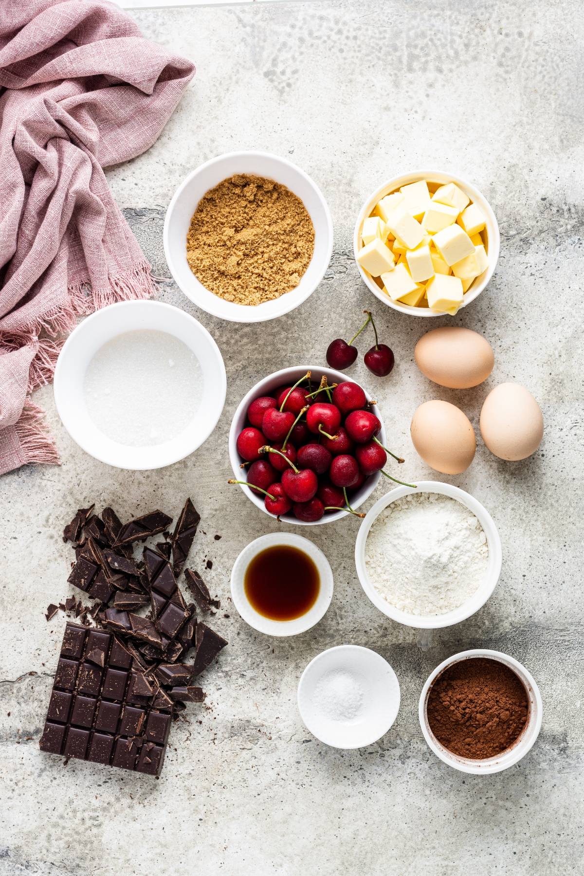 Bowls of baking ingredients—cherries, eggs, flour, sugar, butter, chocolate, cocoa, vanilla, and brown sugar—await your next chocolate and cherry dessert or Double Chocolate Cherry Brownies.