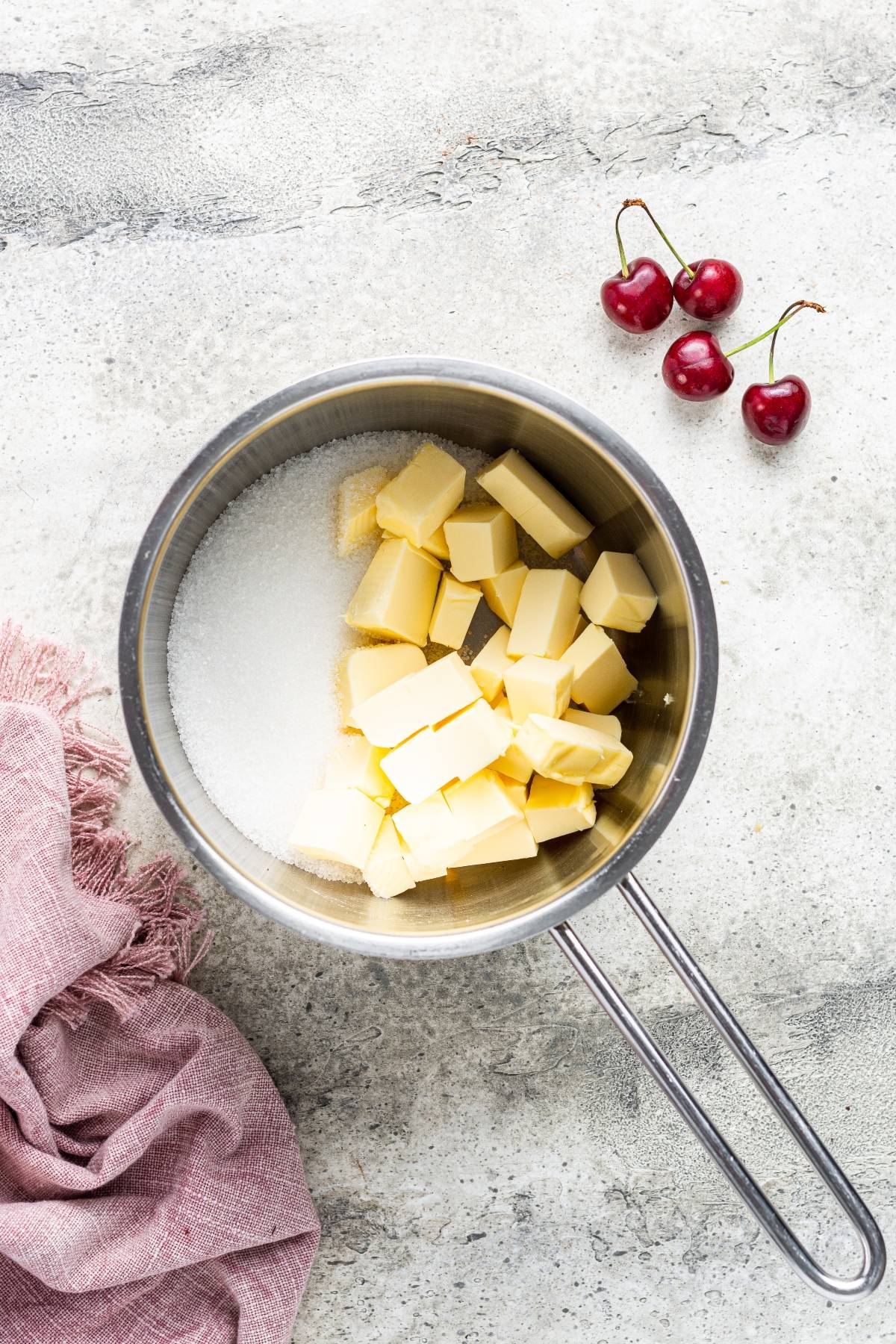 A saucepan with cubed butter and sugar, ideal for making fresh cherry brownies, sits next to three cherries and a pink cloth on a gray surface.