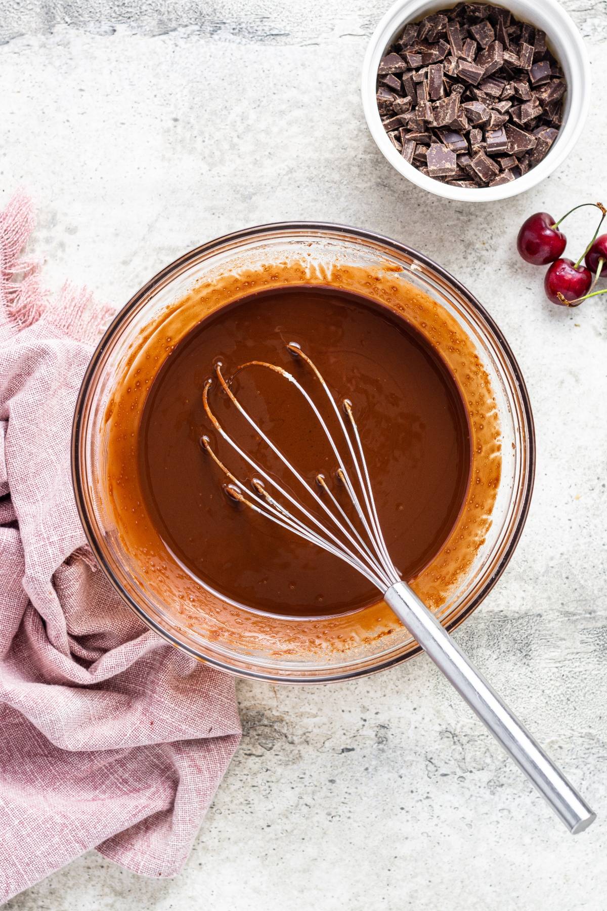 A whisk in a bowl of chocolate batter, with chocolate chunks and cherries nearby on a light surface—perfect for baking decadent Double Chocolate Cherry Brownies.