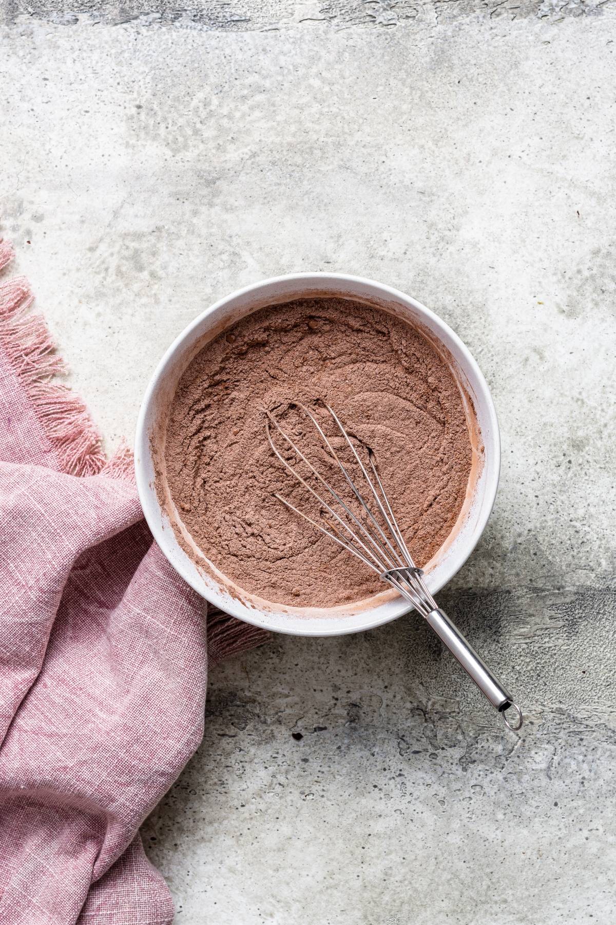 Bowl of cocoa powder mix with a whisk, perfect for Double Chocolate Cherry Brownies, next to a pink cloth on a textured surface.