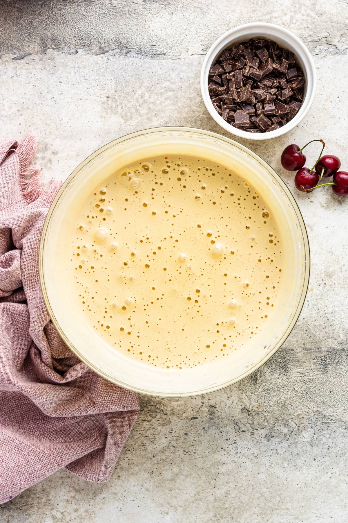 A bowl of frothy batter sits beside chocolate chunks, fresh cherries, and a pink cloth—perfect for a Double Chocolate Cherry Brownies recipe or any delicious chocolate and cherry dessert.