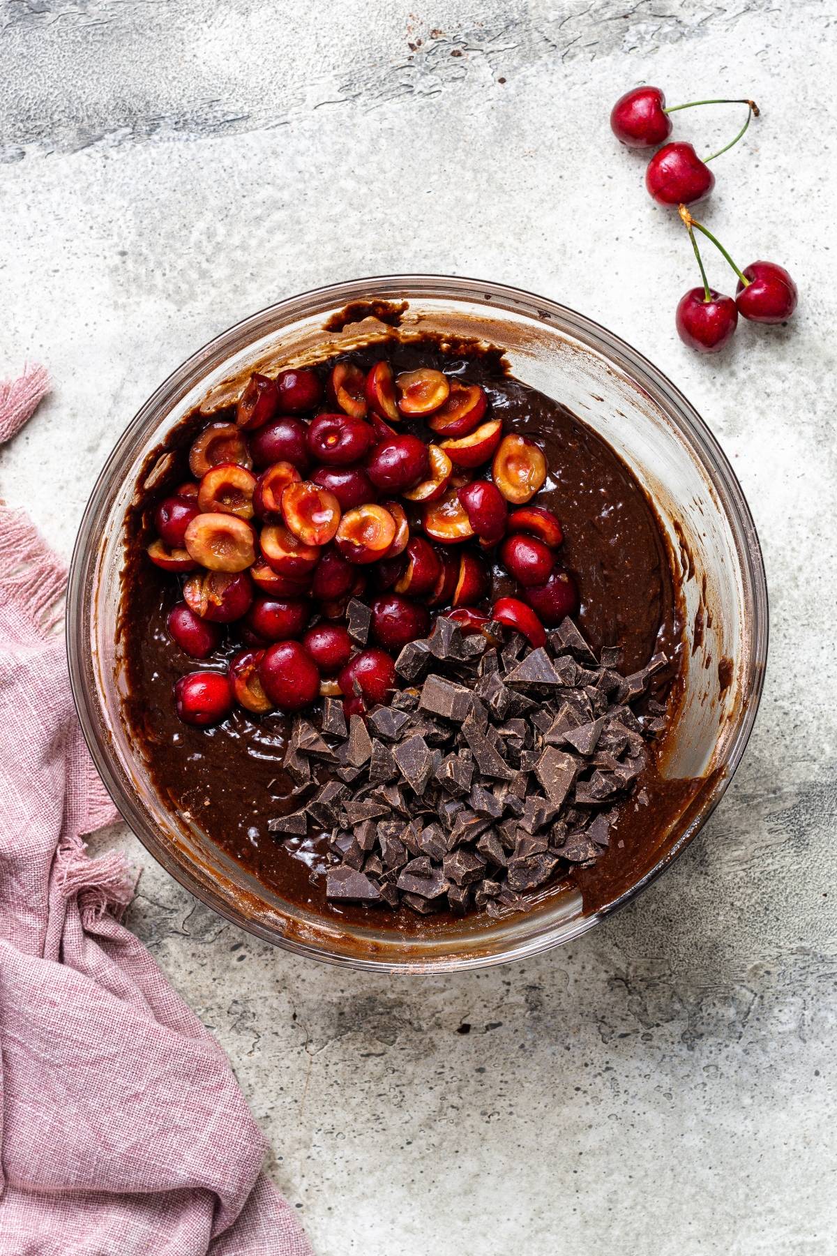 A bowl of brownie batter with chopped cherries and chocolate chunks on top, next to fresh cherries and a pink cloth—an easy cherry dessert perfect for anyone craving a chocolate and cherry dessert.