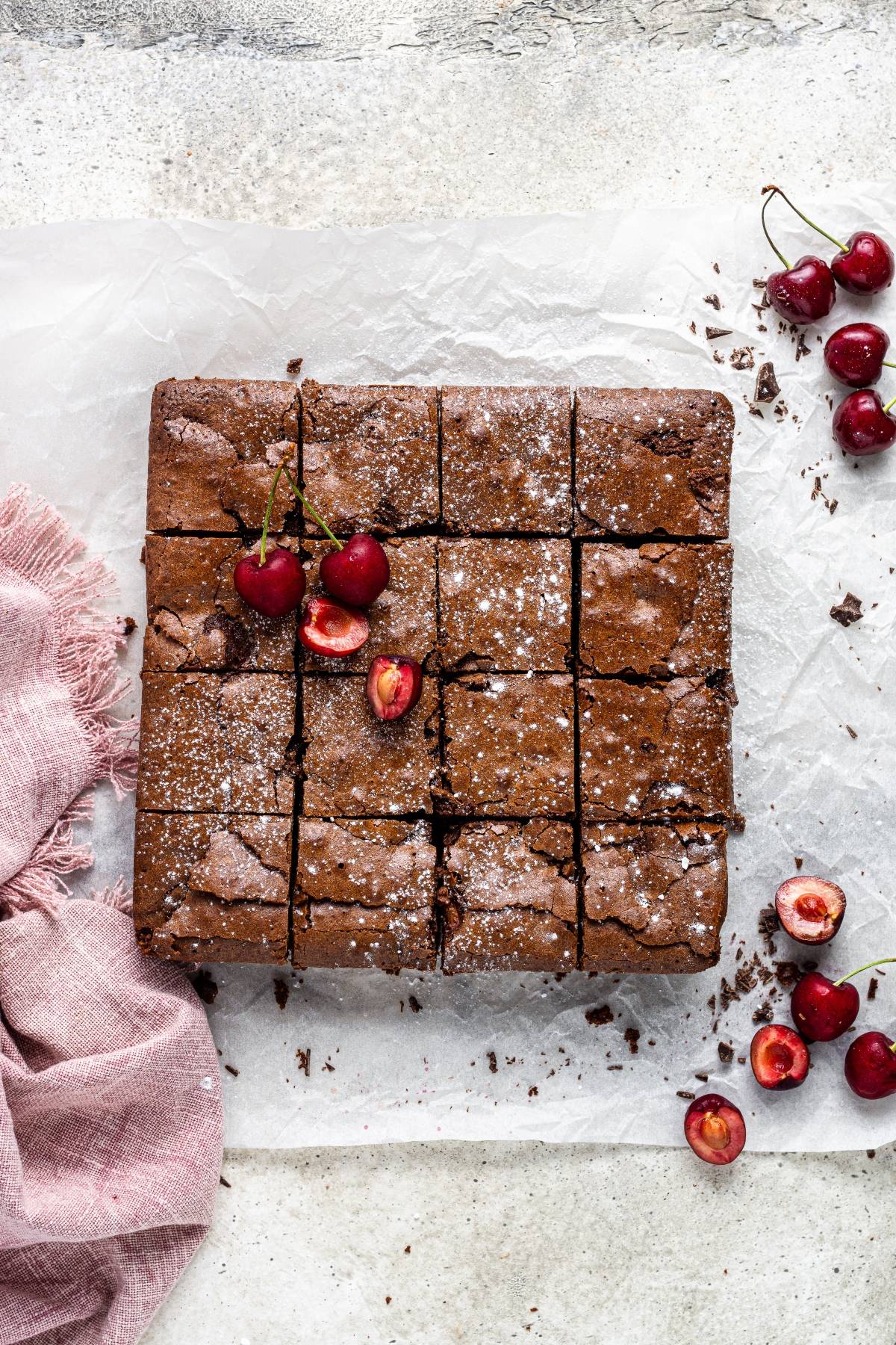 A square chocolate cake, cut into slices, topped with cherries and powdered sugar on parchment paper—an easy cherry dessert perfect for any occasion.