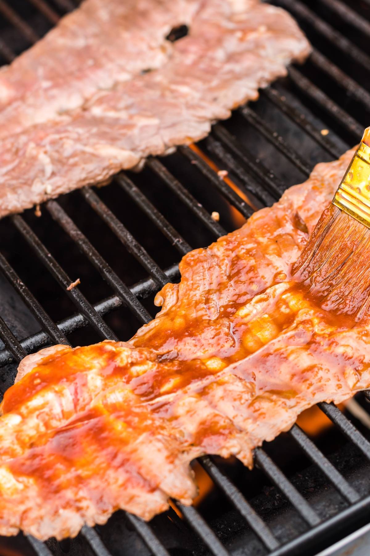 A brush spreads sauce on a thin slice of meat grilling on a barbecue—perfect for a flavorful flank steak recipe or learning how to make carne asada.