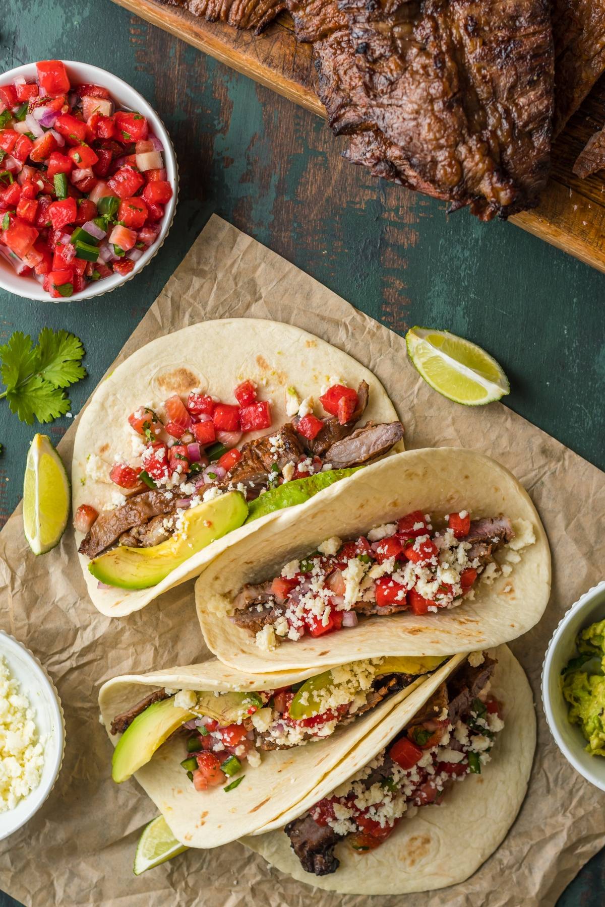 Three flavorful flank steak tacos with avocado, pico de gallo, and cheese on tortillas, featuring the best flank steak marinade. Served with lime wedges and salsa on the side.