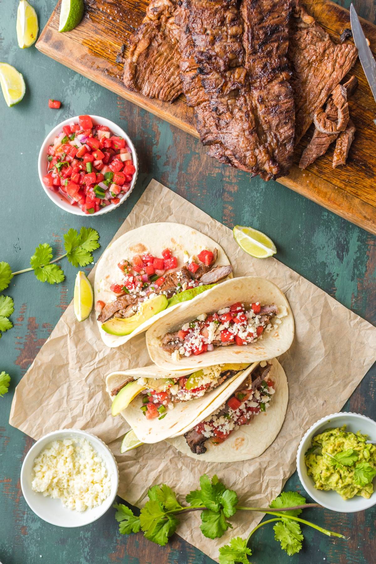 Three flank steak tacos with salsa, cheese, and avocado on parchment, surrounded by steak, guacamole, and garnishes—perfect for showcasing your favorite flank steak recipe.
