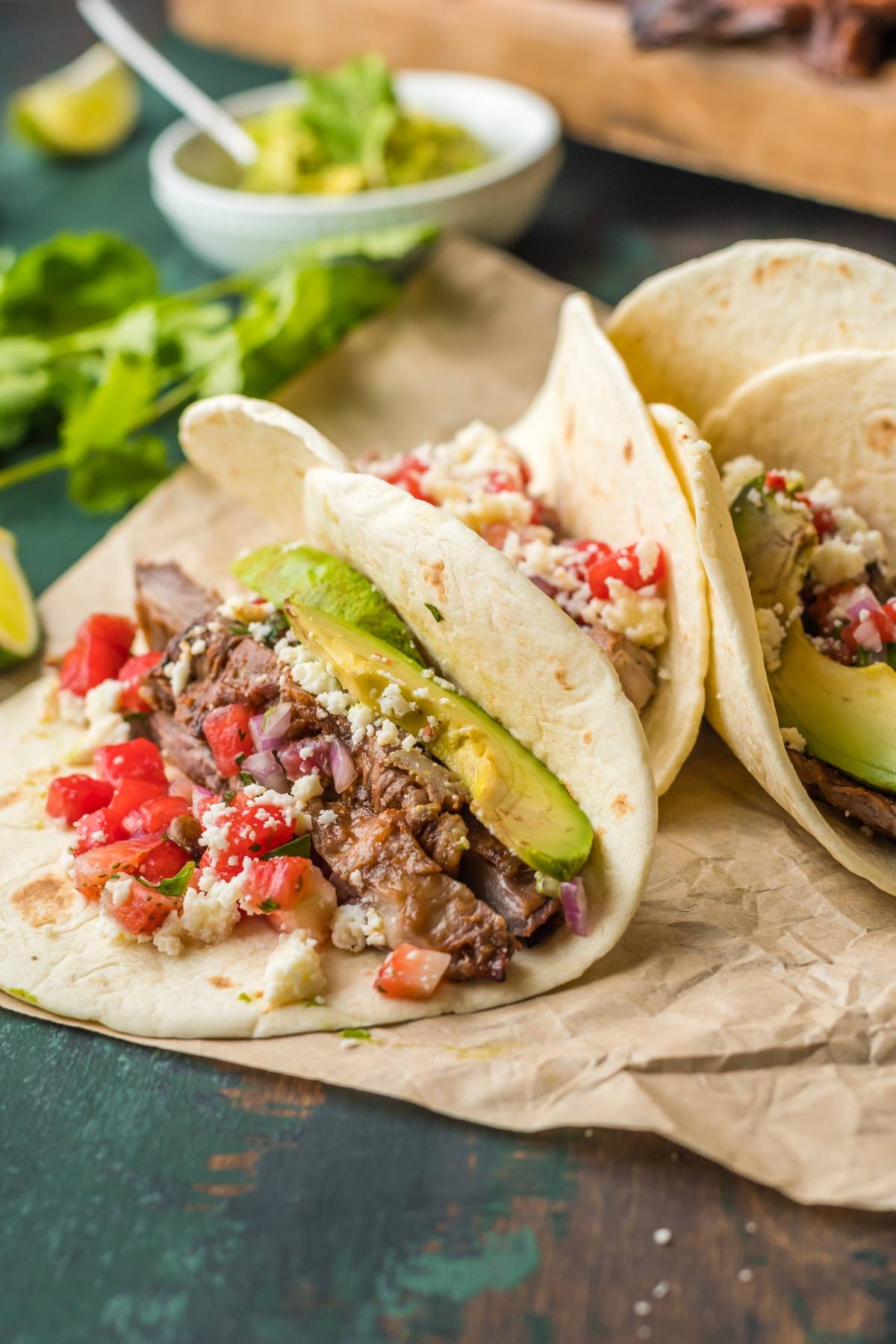 Two soft tacos filled with sliced beef, avocado, tomatoes, cheese, and cilantro on parchment paper—a delicious way to enjoy a classic flank steak recipe.