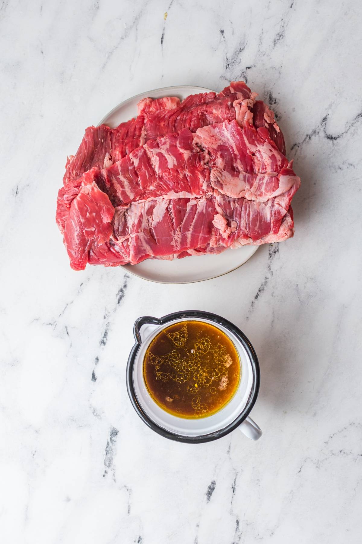 Raw beef on a plate next to a cup of brown liquid marinade on a white marble surface—perfect for trying the best flank steak marinade or learning how to make carne asada.