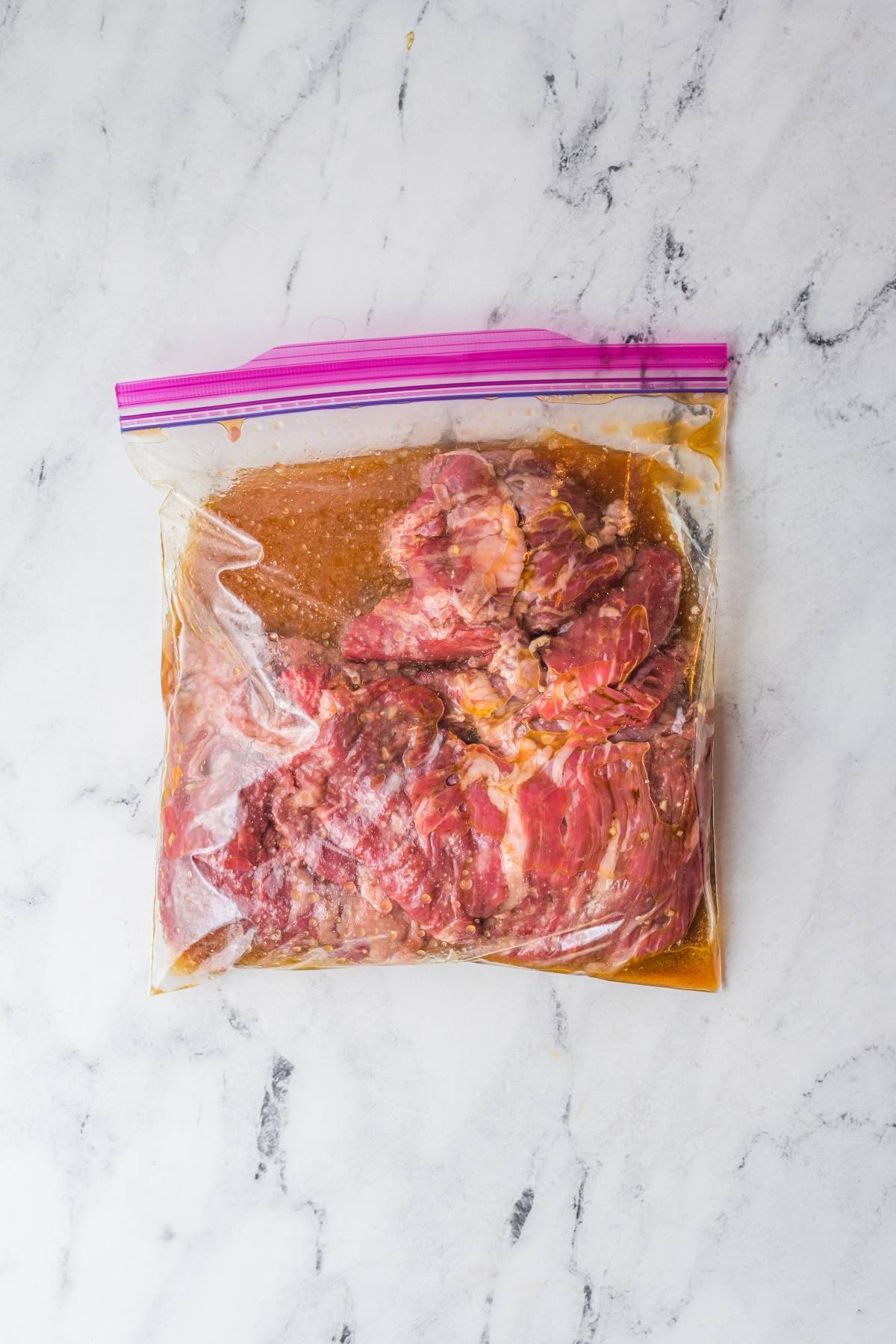 Raw marinated steak in a clear ziplock bag on a white marble surface, ready to become the star of your next flank steak tacos or any top-rated Flank steak recipe.