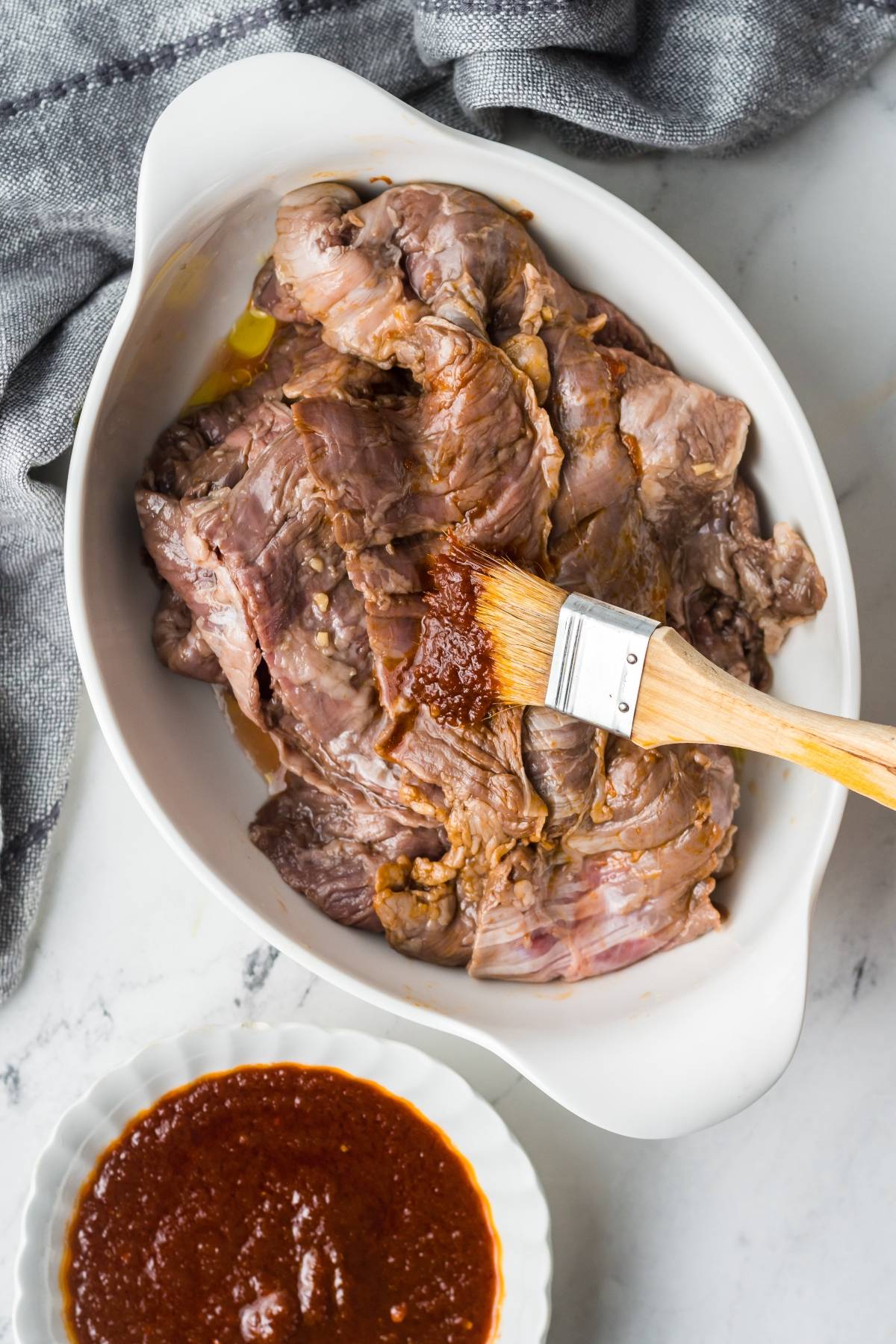 Raw meat in a white dish is being brushed with a vibrant carne asada marinade; a sauce bowl sits nearby on the white countertop.