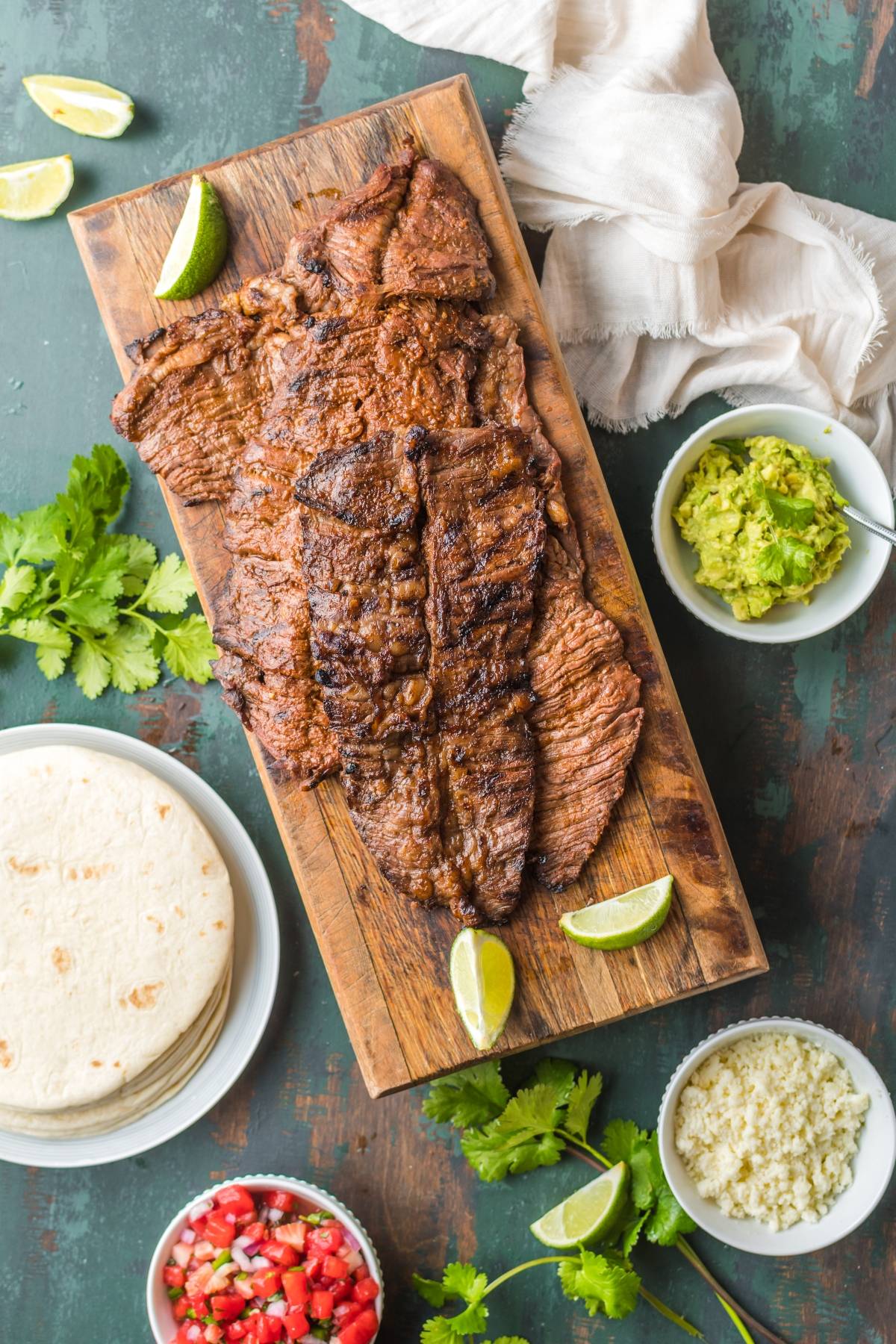 Grilled steak on a wooden board with lime wedges, tortillas, guacamole, salsa, cheese, and cilantro nearby—a delicious spread inspired by the best flank steak marinade.