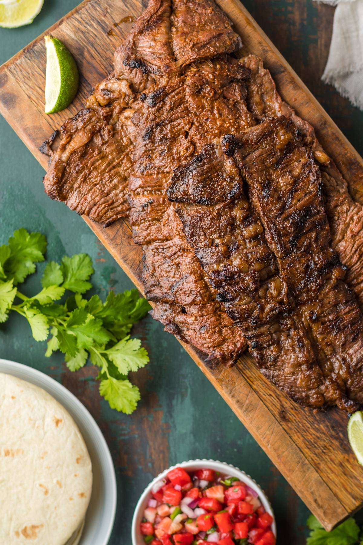 Grilled carne asada steak on a wooden board with lime wedges, cilantro, tortillas, and salsa on the side—the perfect flank steak recipe for delicious tacos and gatherings.