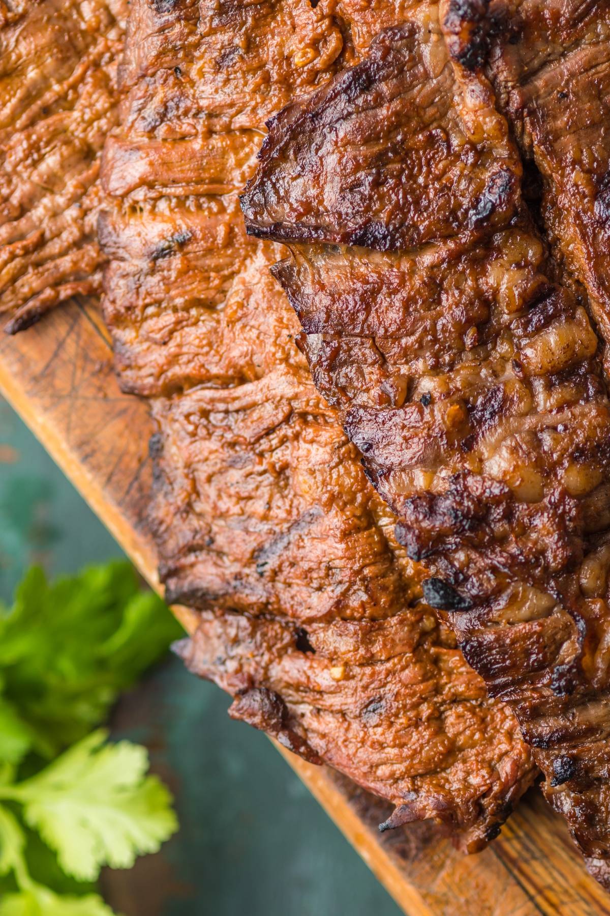 Sliced grilled steak on a wooden board with fresh herbs in the background—perfect for your next flank steak recipe or flavorful carne asada marinade.