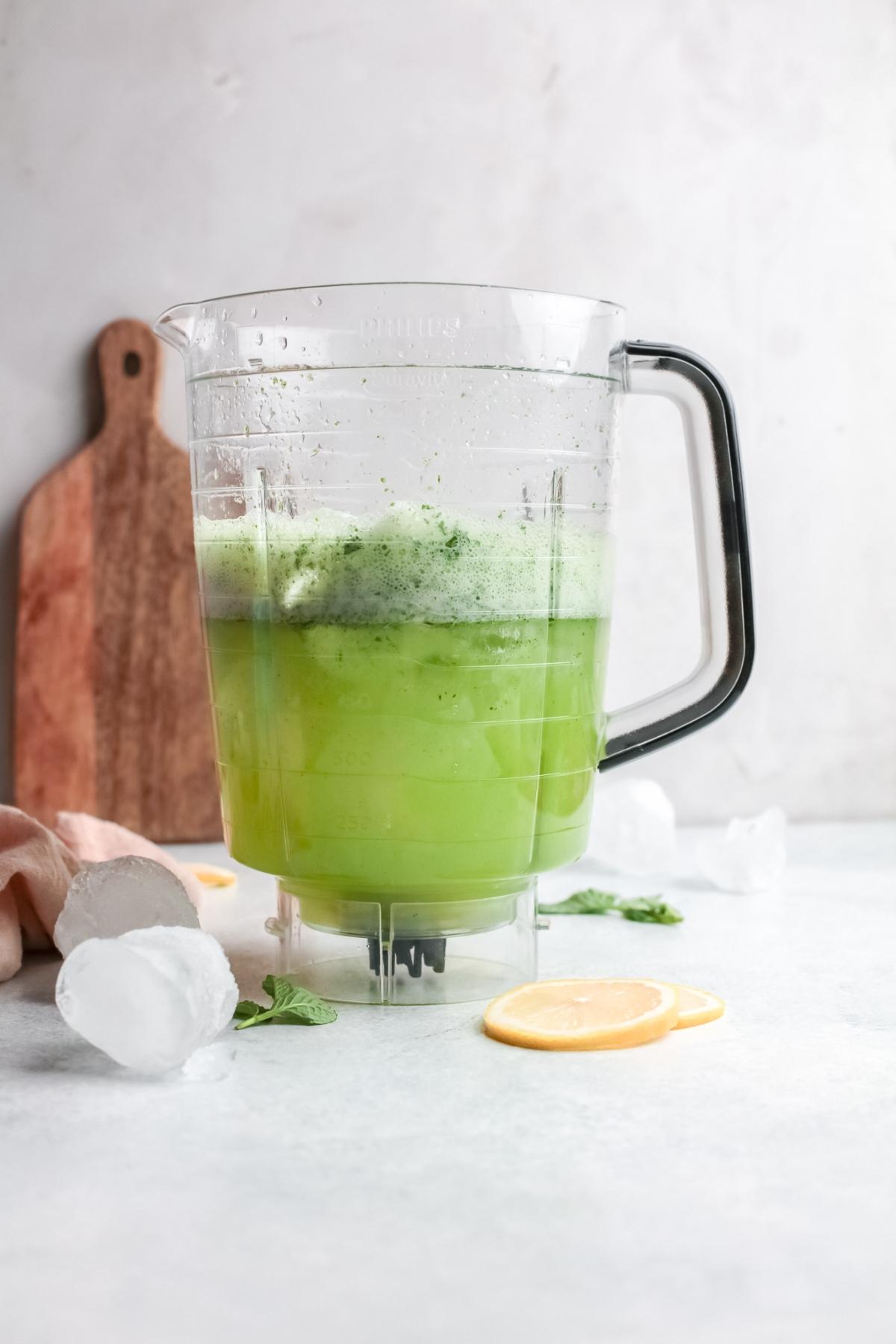 A blender filled with green juice, ice cubes, lemon slices, and mint leaves on a light surface, perfect for making a refreshing limonana mint slush drink.