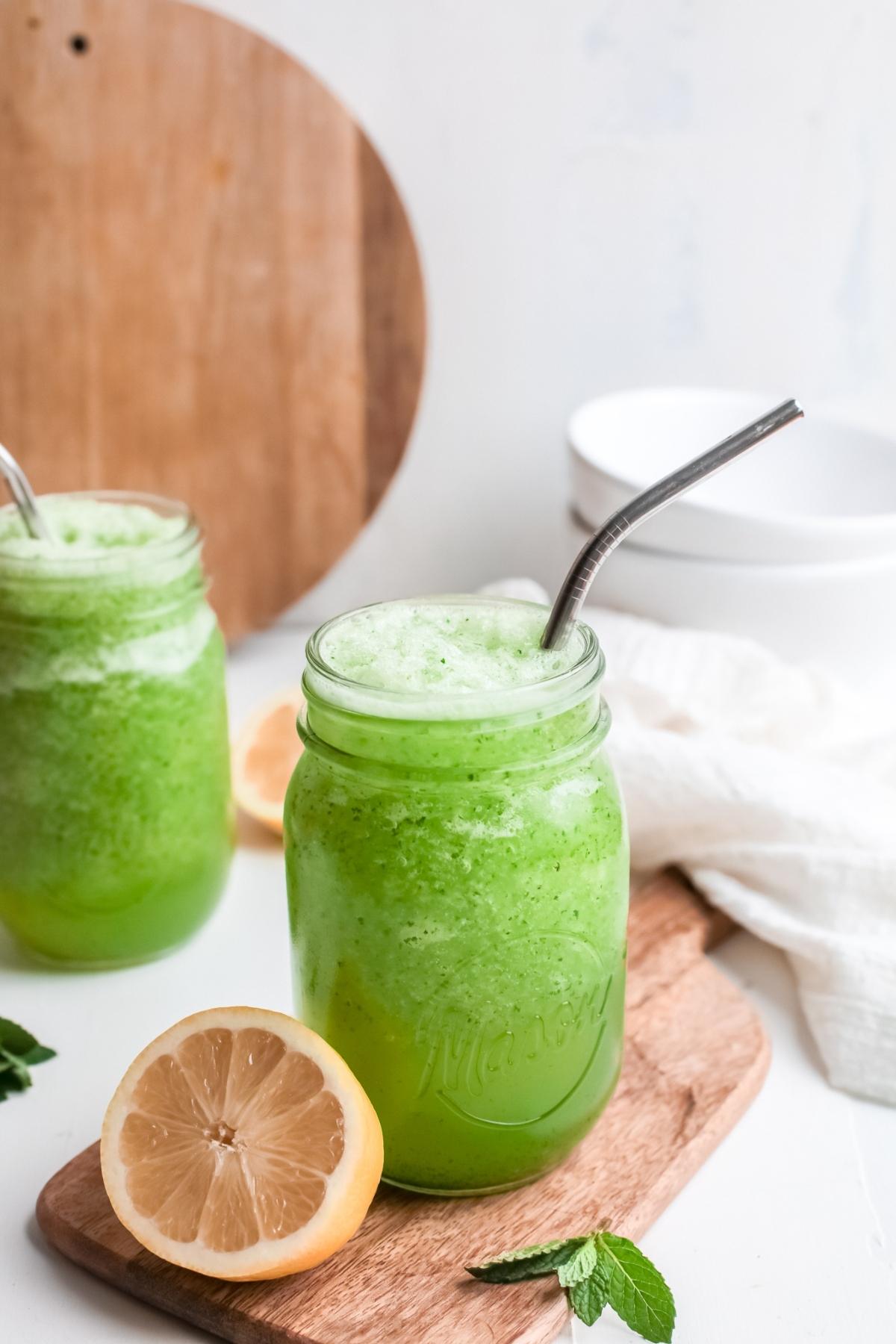 Two mason jars of frothy green lemon and mint drink with metal straws, mint leaves, and a halved lemon on a wooden board.