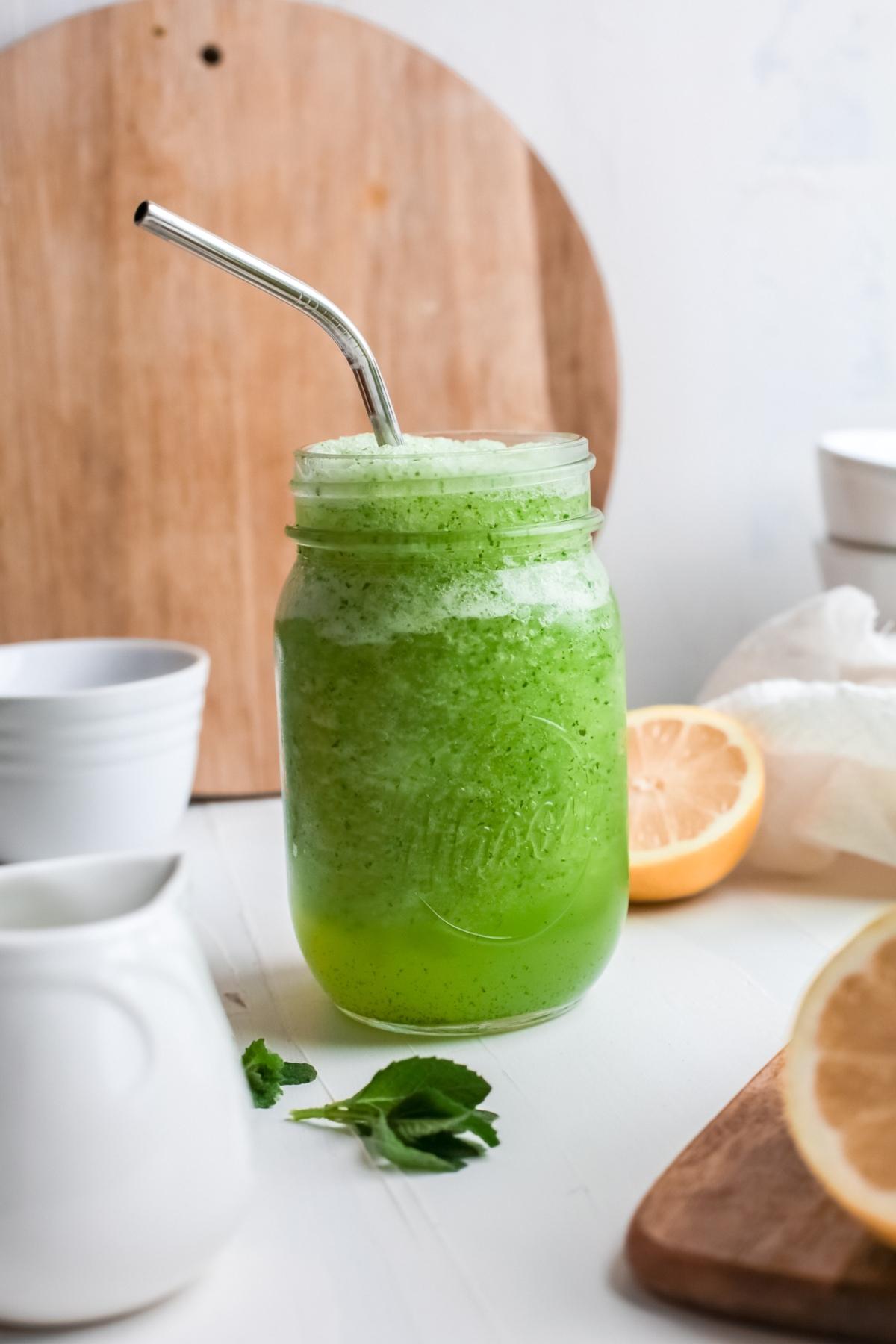 A mason jar of green juice with foam and a metal straw, reminiscent of a refreshing lemon and mint drink, sits next to lemons and cups on a white table.
