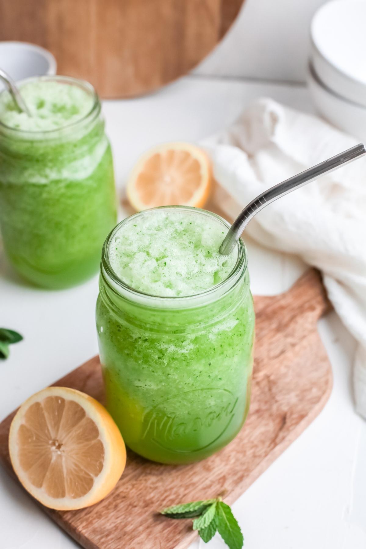 A mason jar of Frozen Mint Lemonade - limonana with a metal straw, next to lemon halves and mint leaves on a wooden board.