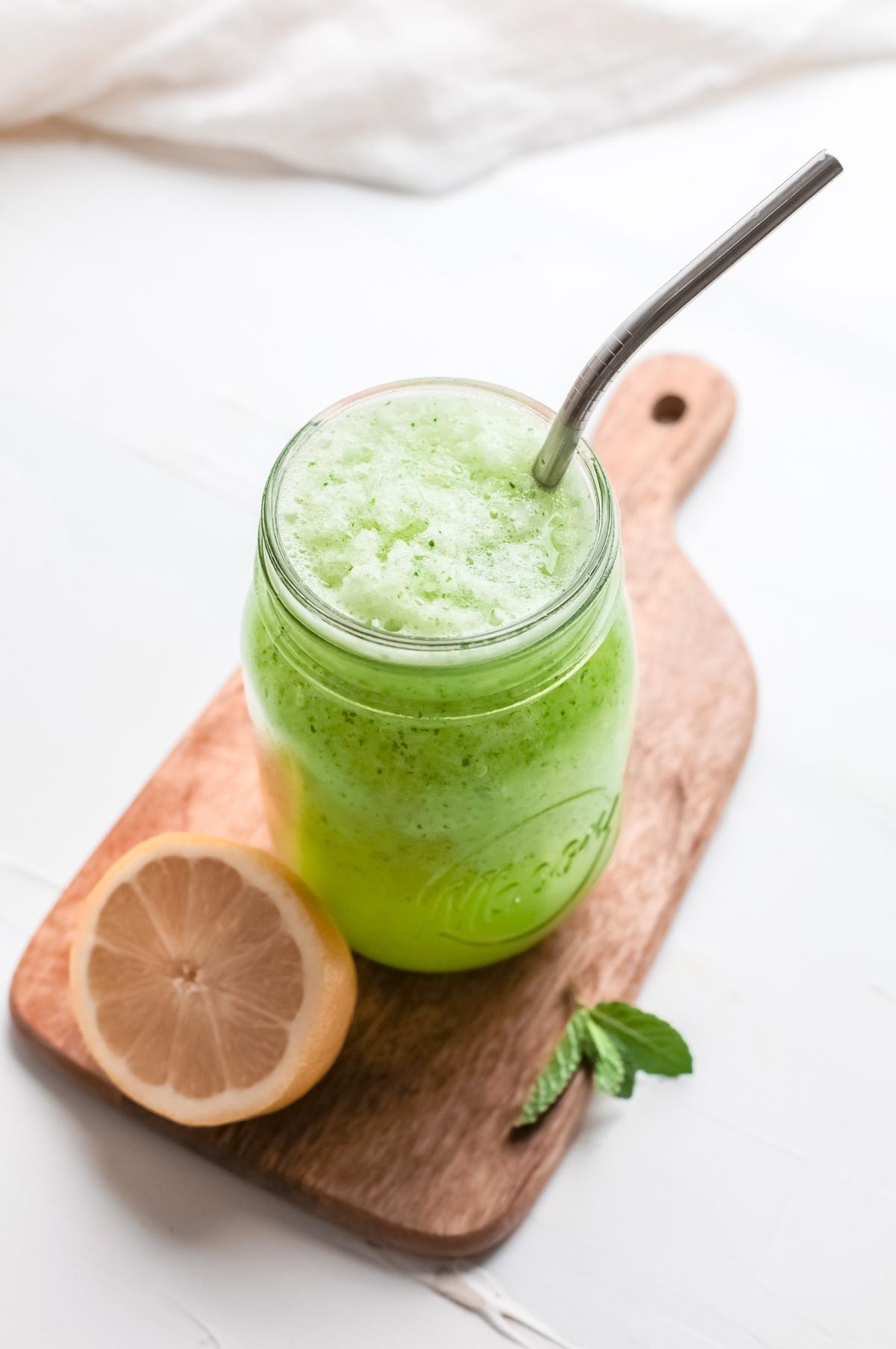 A green mint slush drink in a mason jar with a metal straw, beside a lemon half and fresh mint on a wooden board.