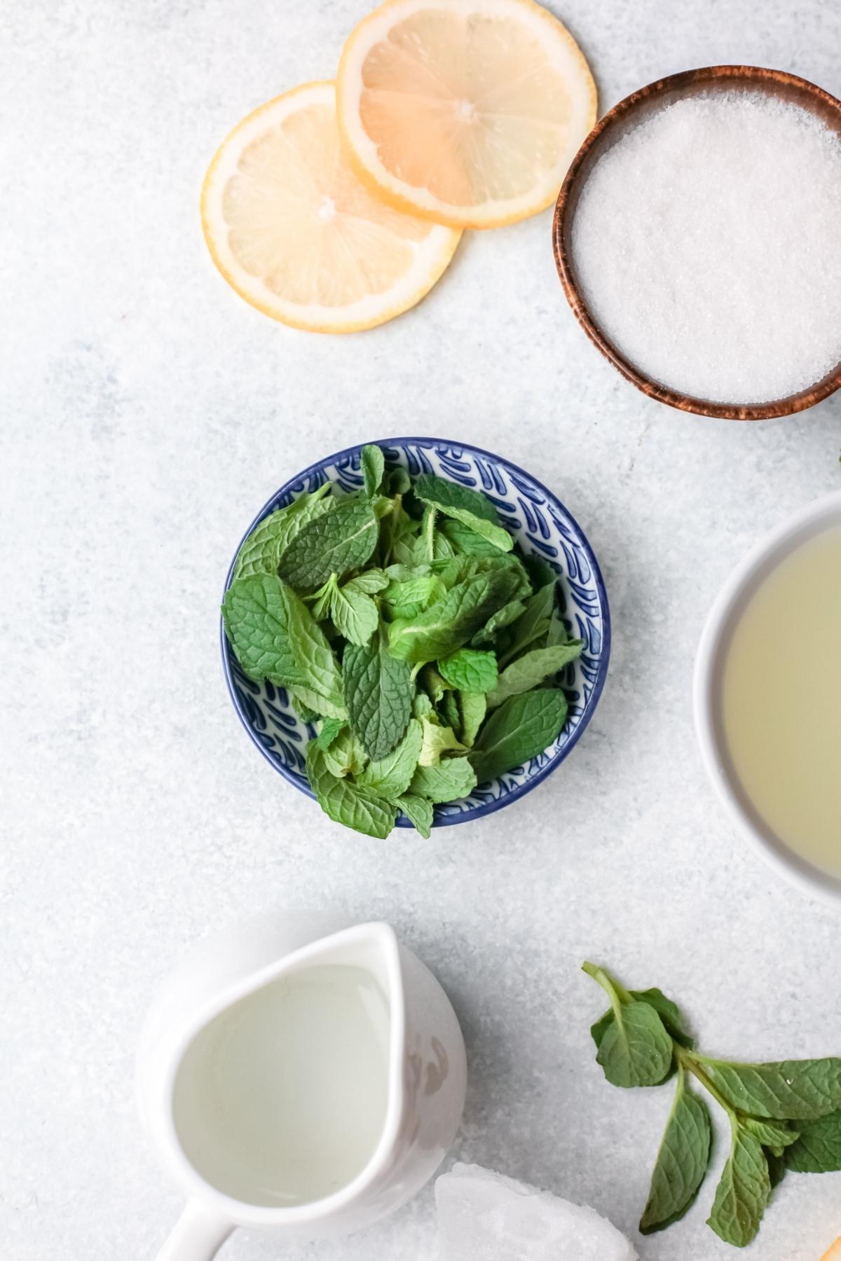 Top view of mint leaves in a bowl, lemon slices, sugar, lemon juice, and a small pitcher on a light surface—perfect ingredients for a refreshing frozen lemonade recipe or classic lemon and mint drink.